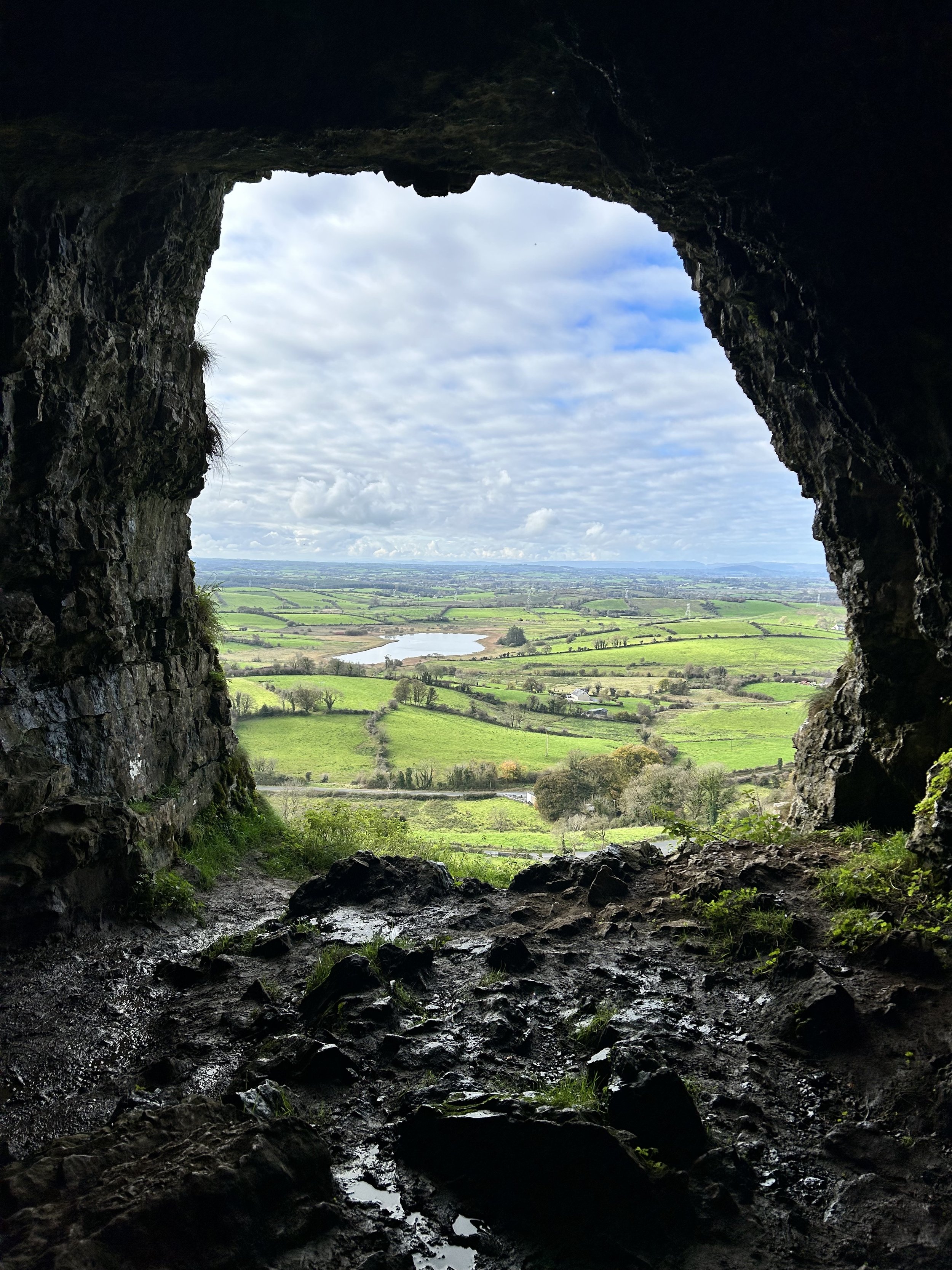 View from inside a cave looking out over a scenic landscape with green fields and cloudy sky.