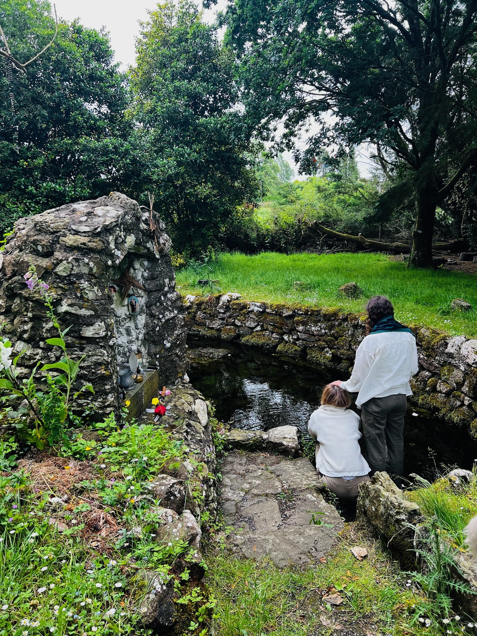Stone well surrounded by greenery with people kneeling near the water.