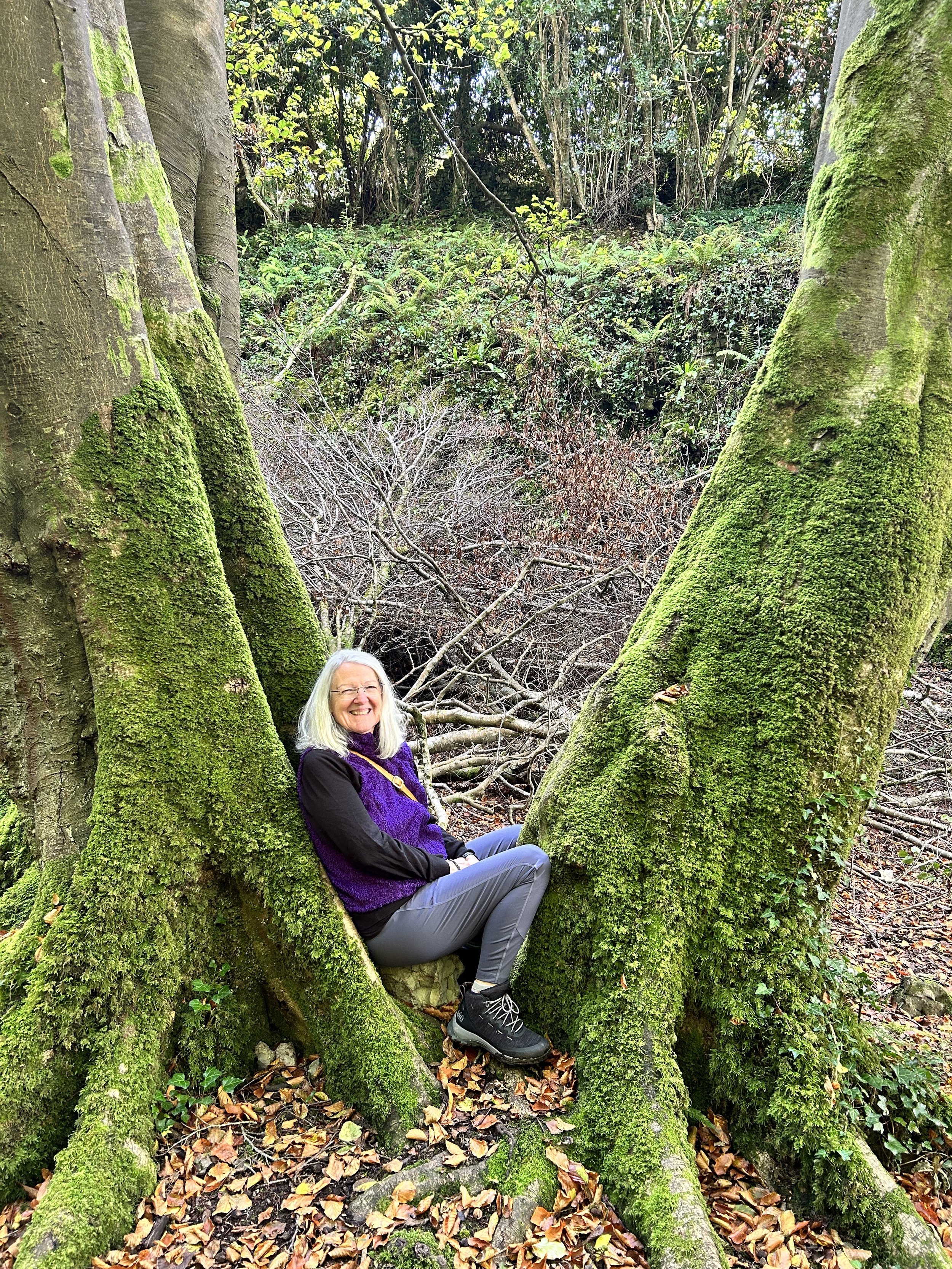 Woman with white hair sitting between moss-covered tree trunks in a forest.