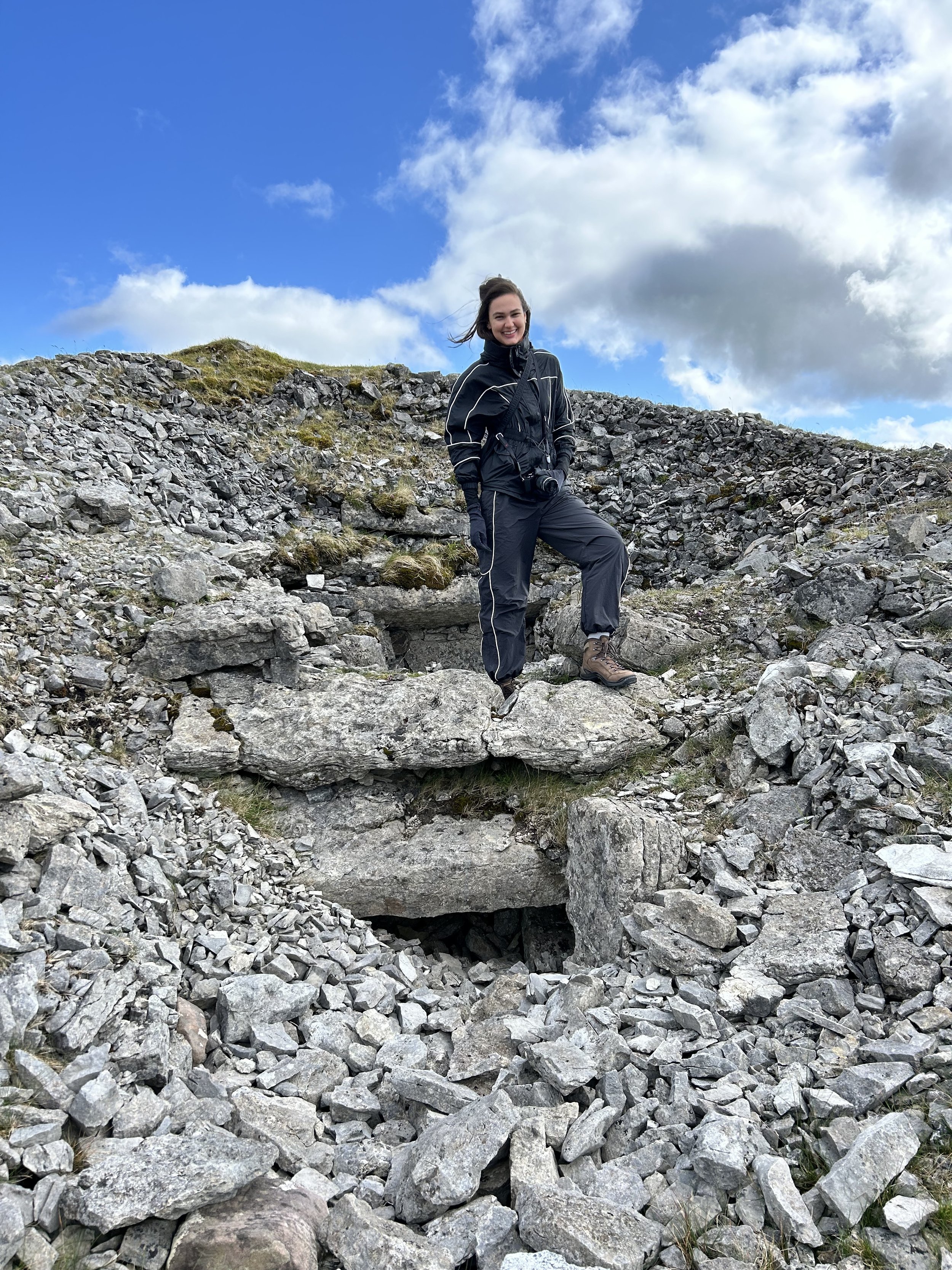 Person in hiking gear standing on rocky terrain with blue sky and clouds.
