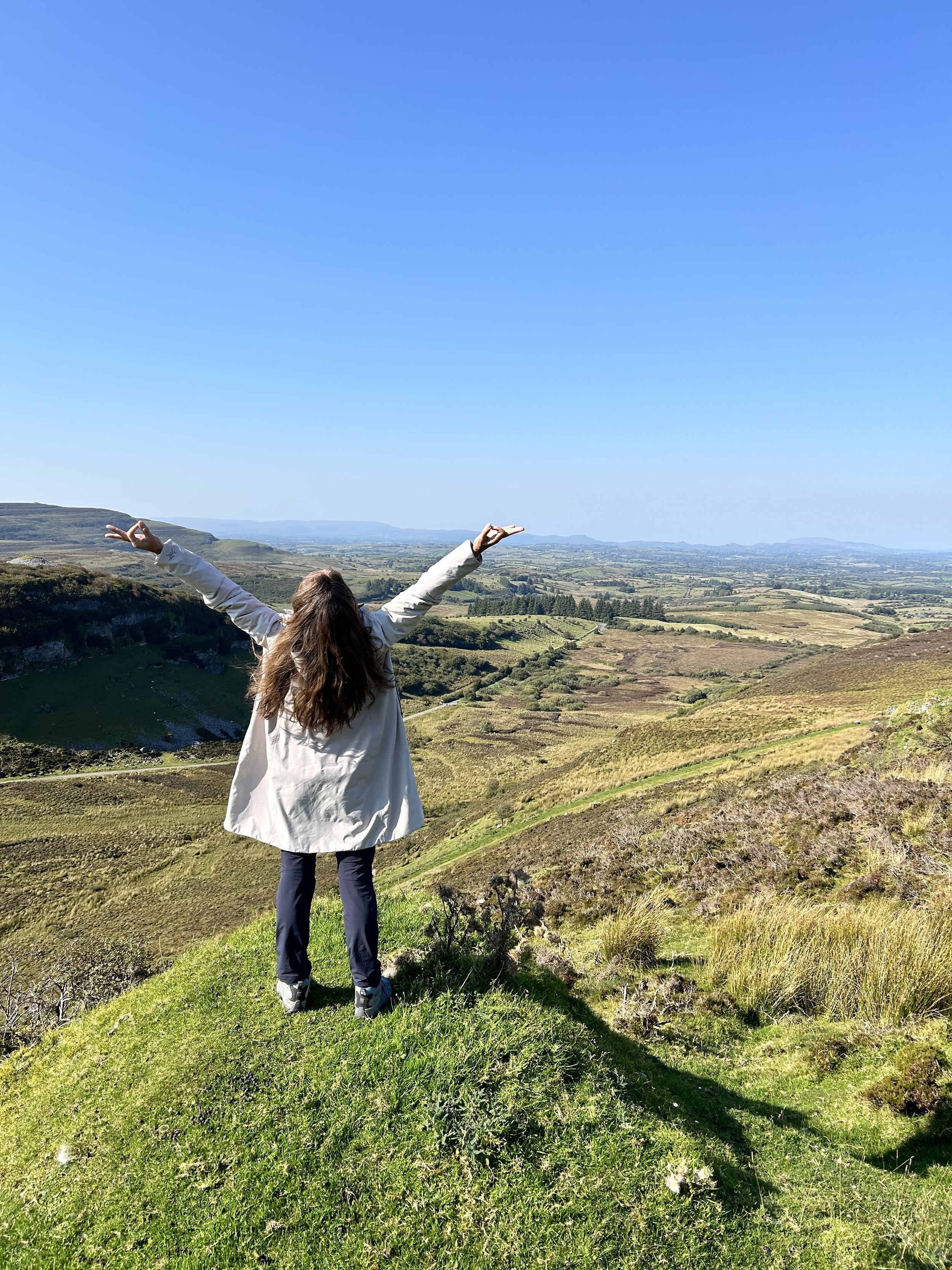 Person standing on a grassy hill with arms raised, overlooking a vast landscape of rolling hills and fields under a clear blue sky.