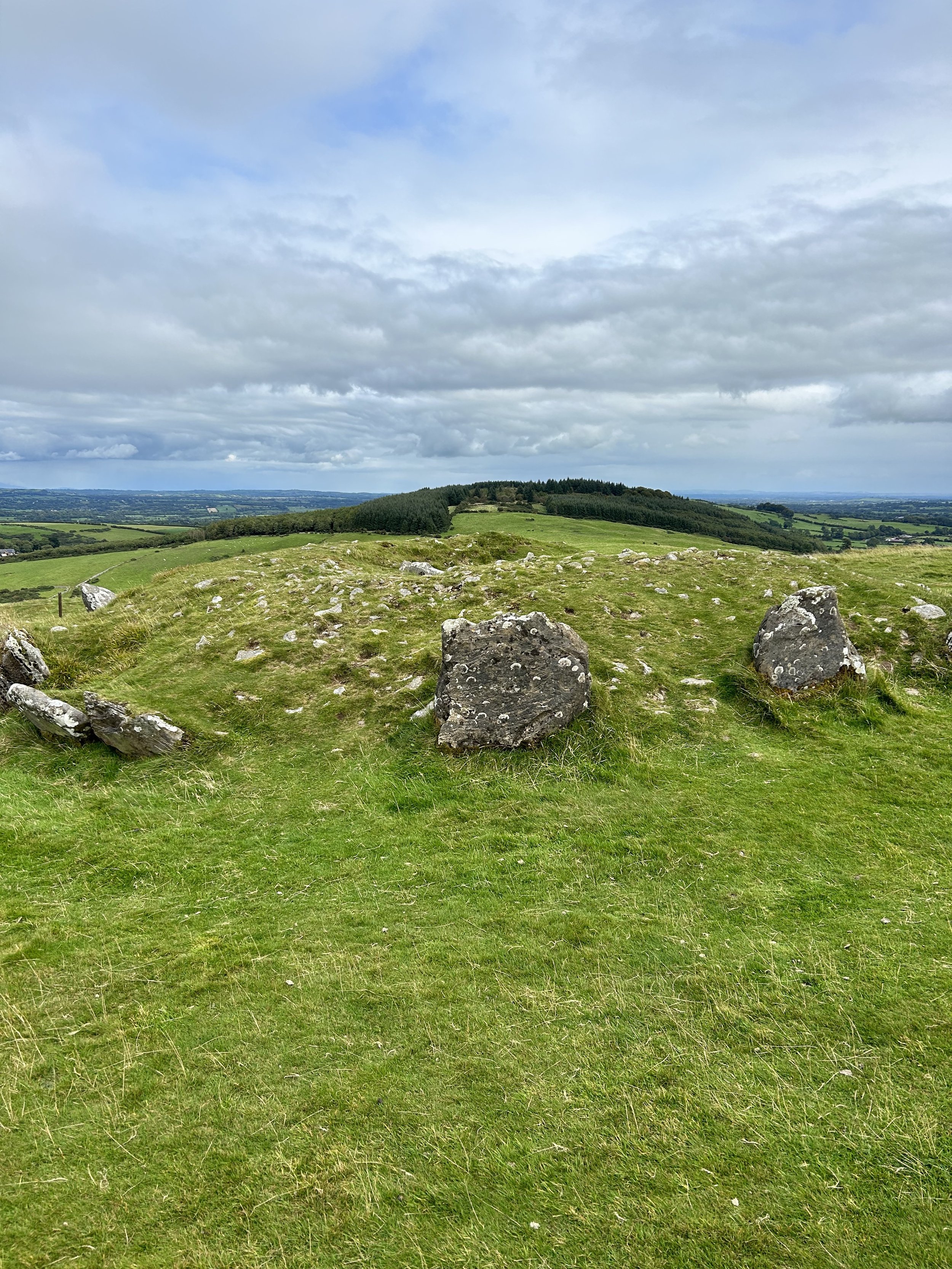 Grass-covered hill with large rocks and a view of distant hills and cloudy sky.
