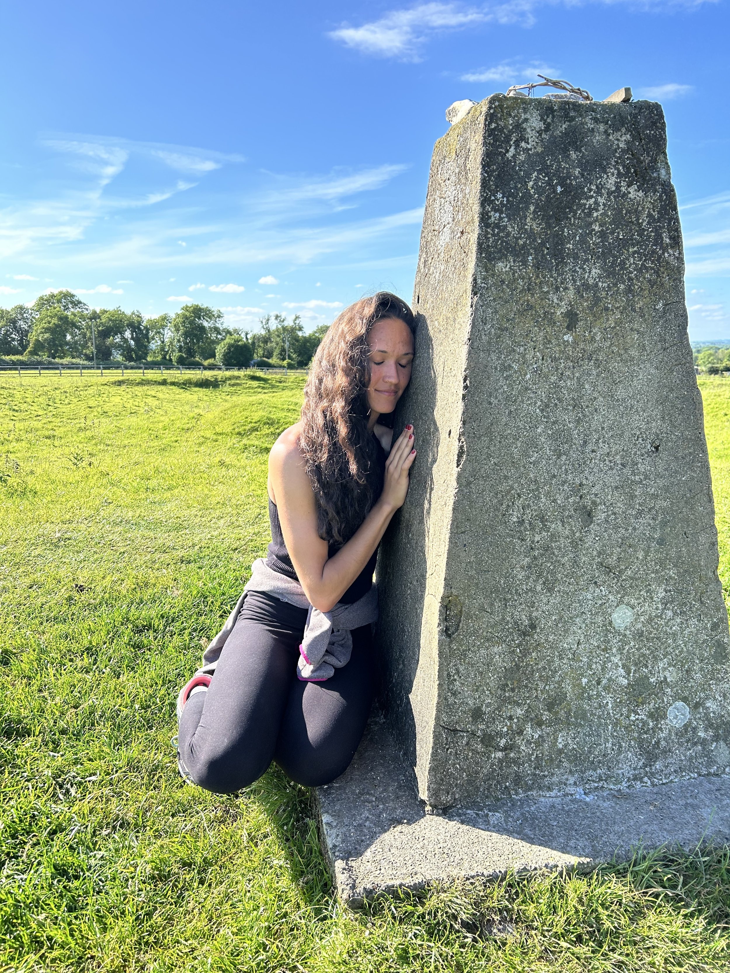 Person kneeling and resting head against a large stone pillar in a grassy field with a clear blue sky.
