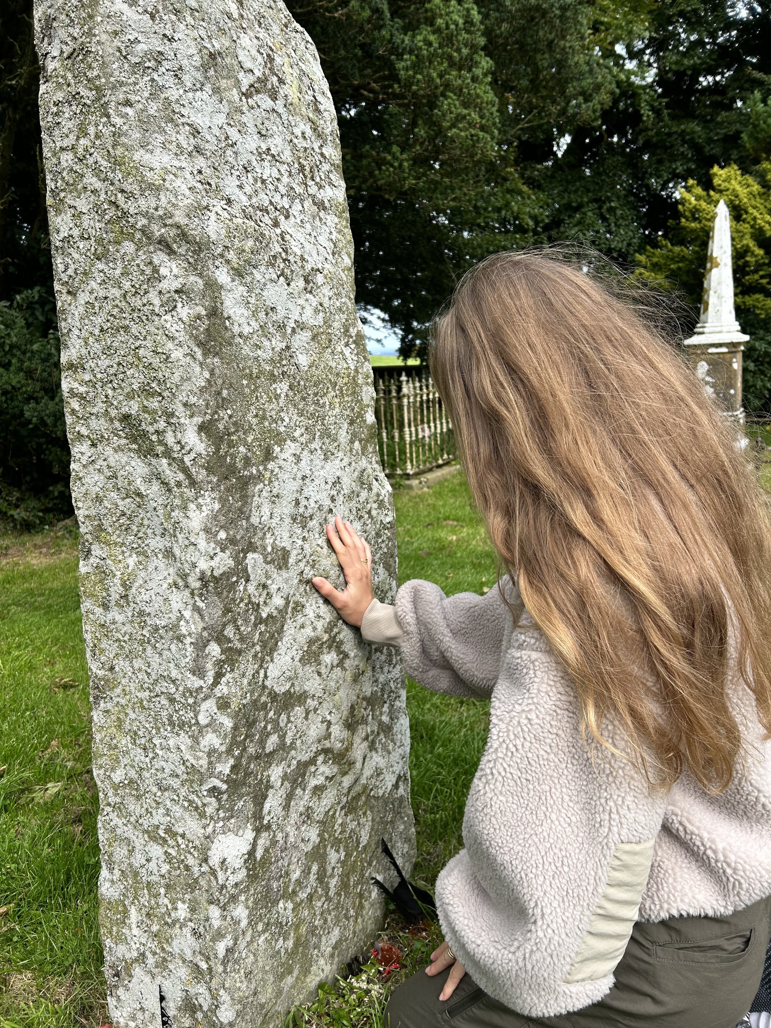 Person touching a large, ancient stone slab with weathered texture in a grassy outdoor setting.