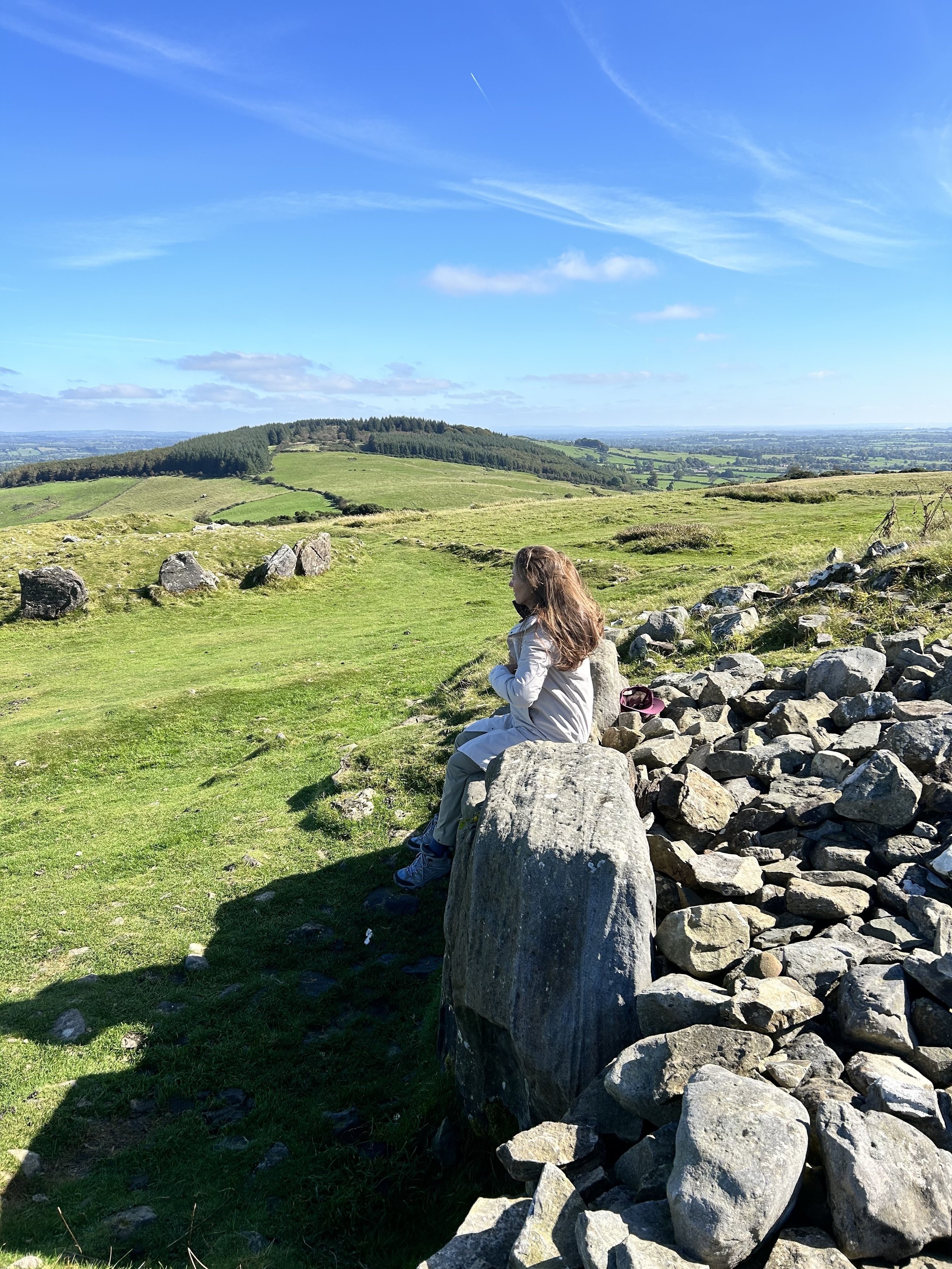 A person sitting on a large rock amidst a grassy landscape with hills and scattered rocks, under a clear blue sky.