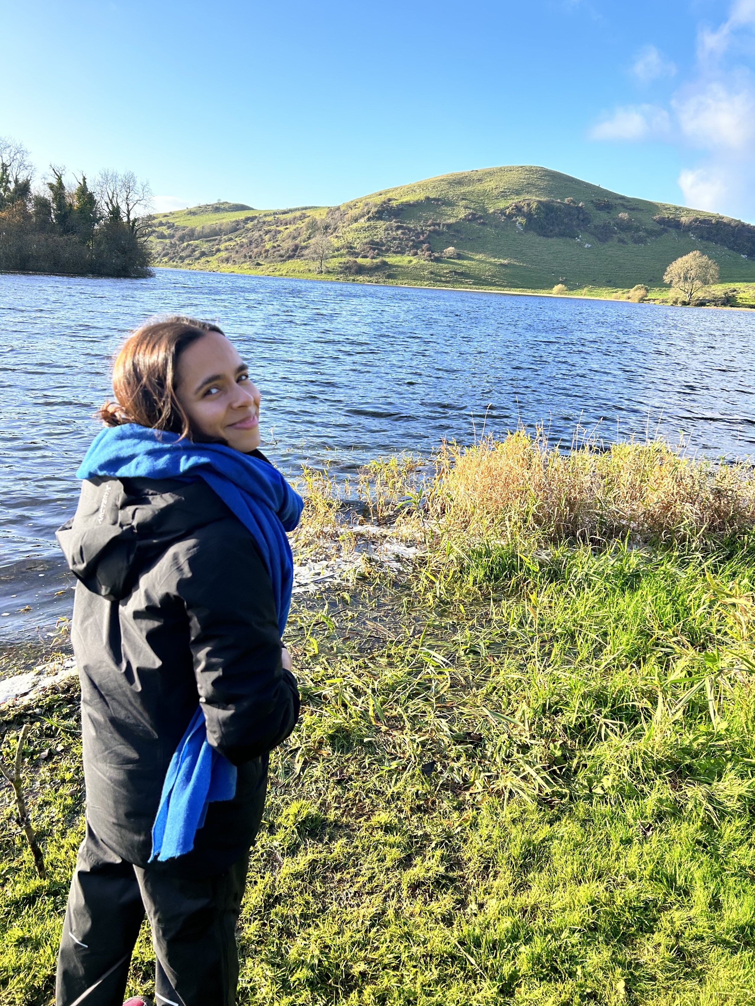 Person wearing a black jacket and blue scarf standing by a lake with green hills in the background on a sunny day.