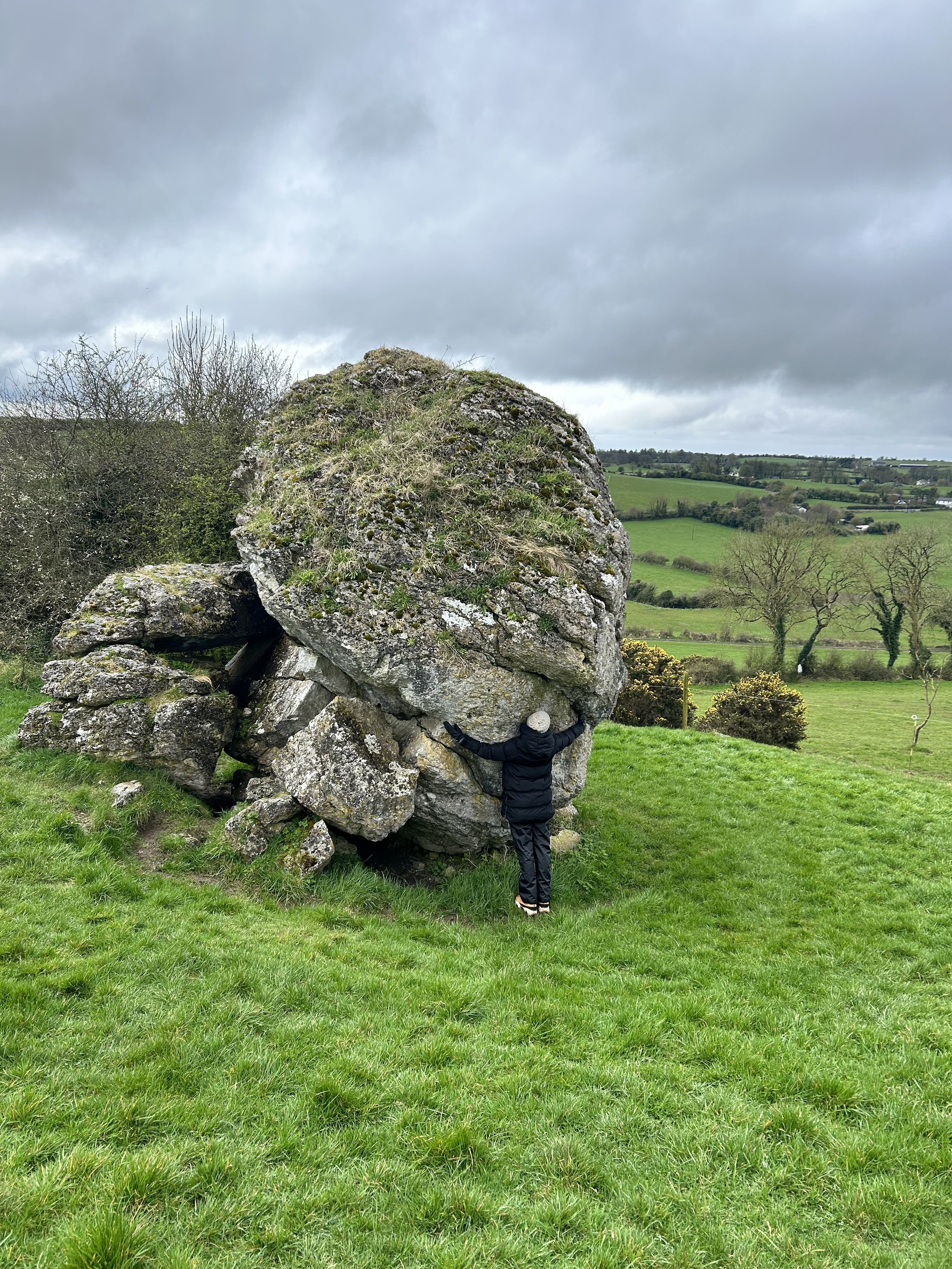 Person standing against a large boulder in a grassy field, with a cloudy sky in the background.
