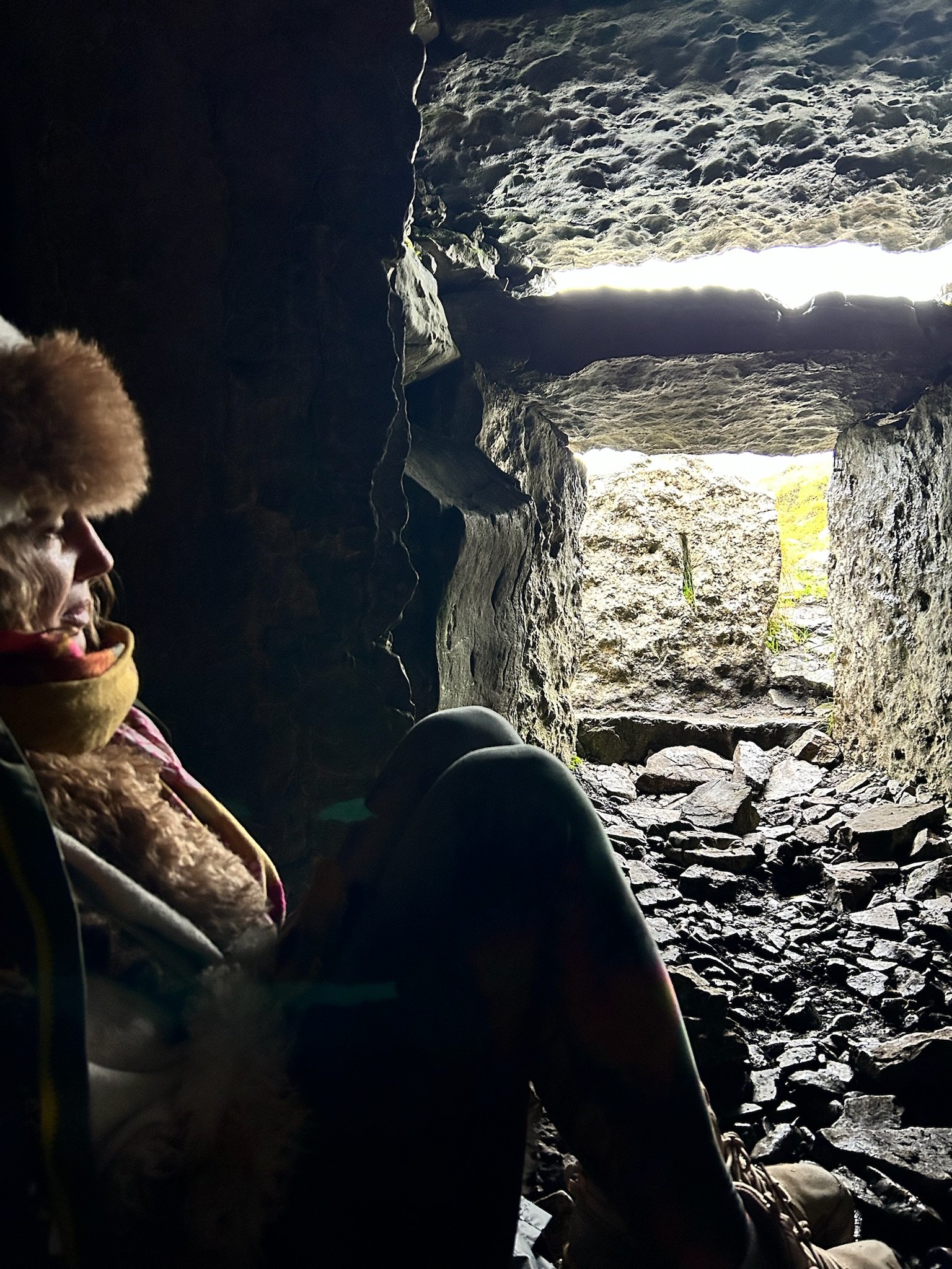 Person wearing winter clothing sitting inside a rocky cave with light streaming in from the entrance.