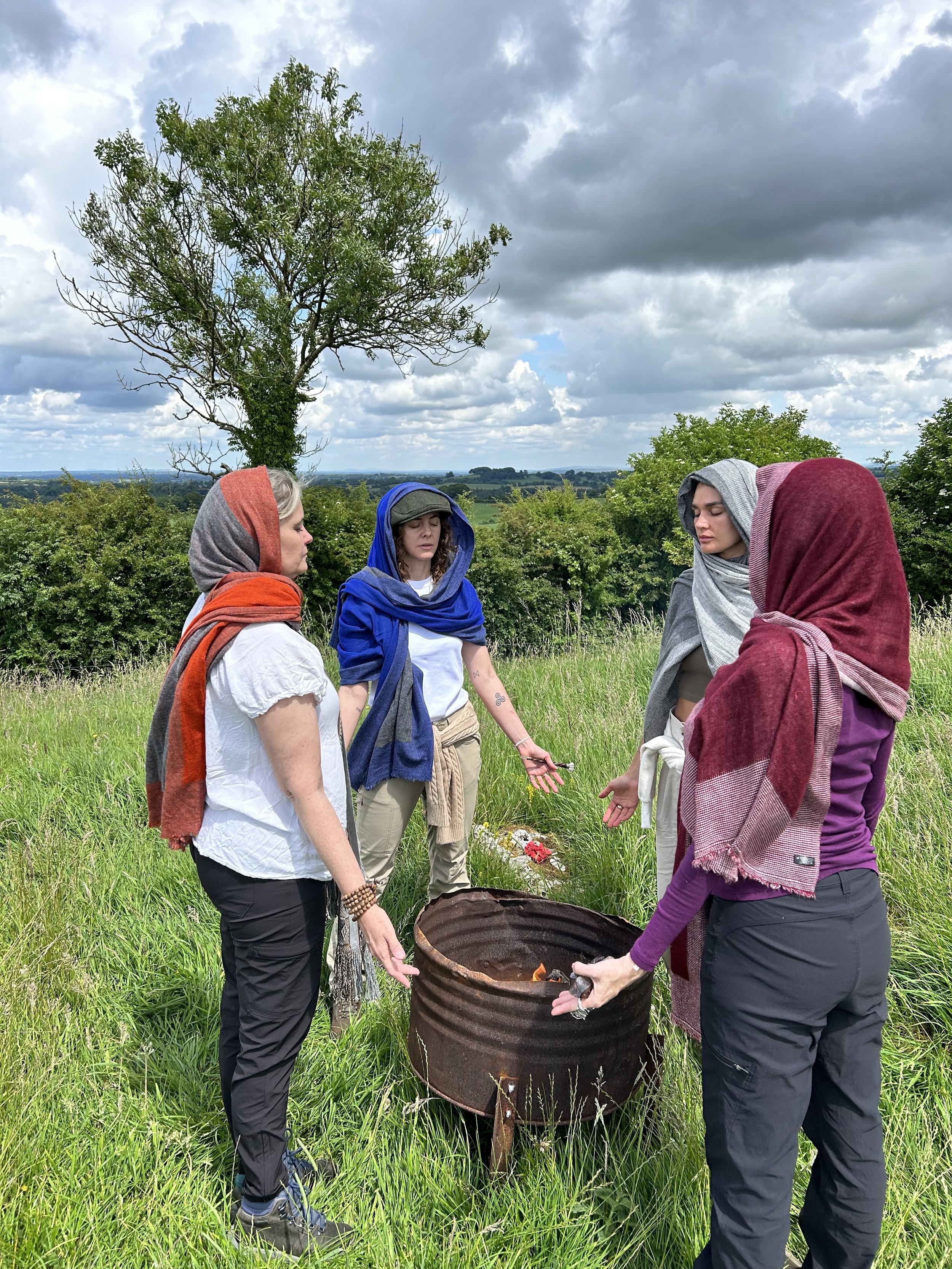 Four people wearing colorful scarves standing in a circle around a rusted metal fire pit in a grassy field, with a tree and clouds in the background.