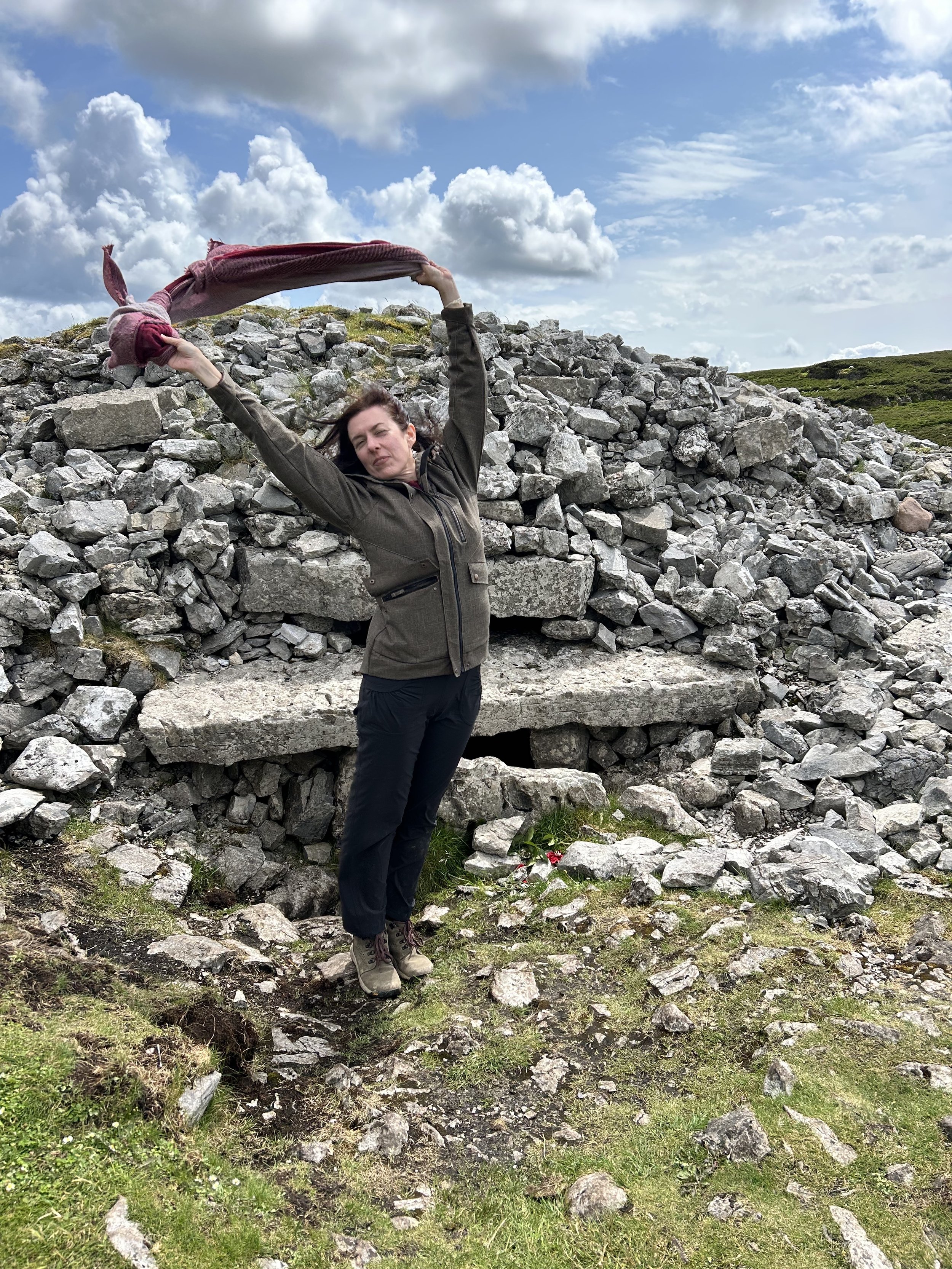 Person holding a scarf in the air, standing in front of a stone cairn under a cloudy sky.