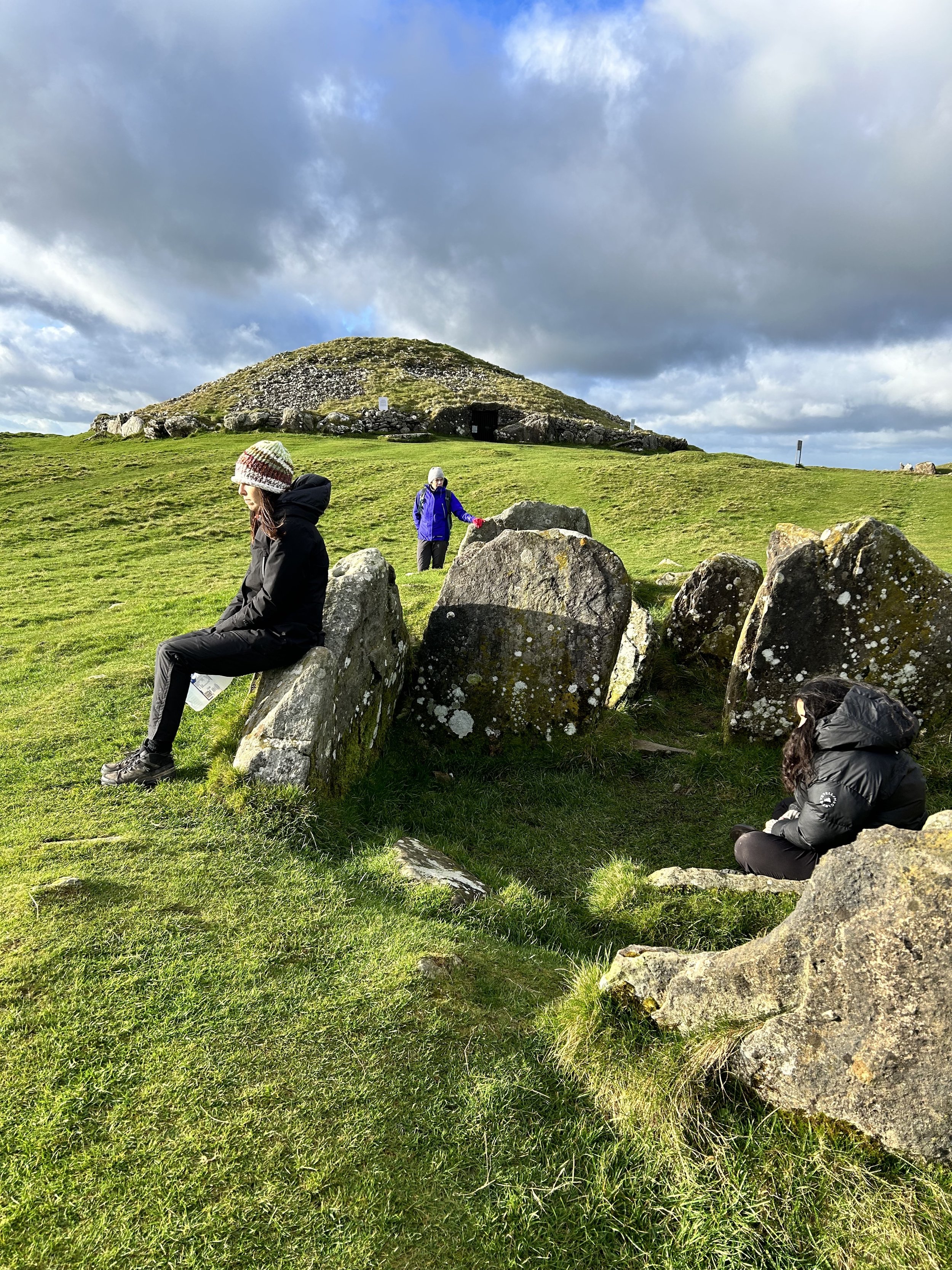 People sitting on and near ancient stone formations with a grassy hill and mound in the background under a cloudy sky.