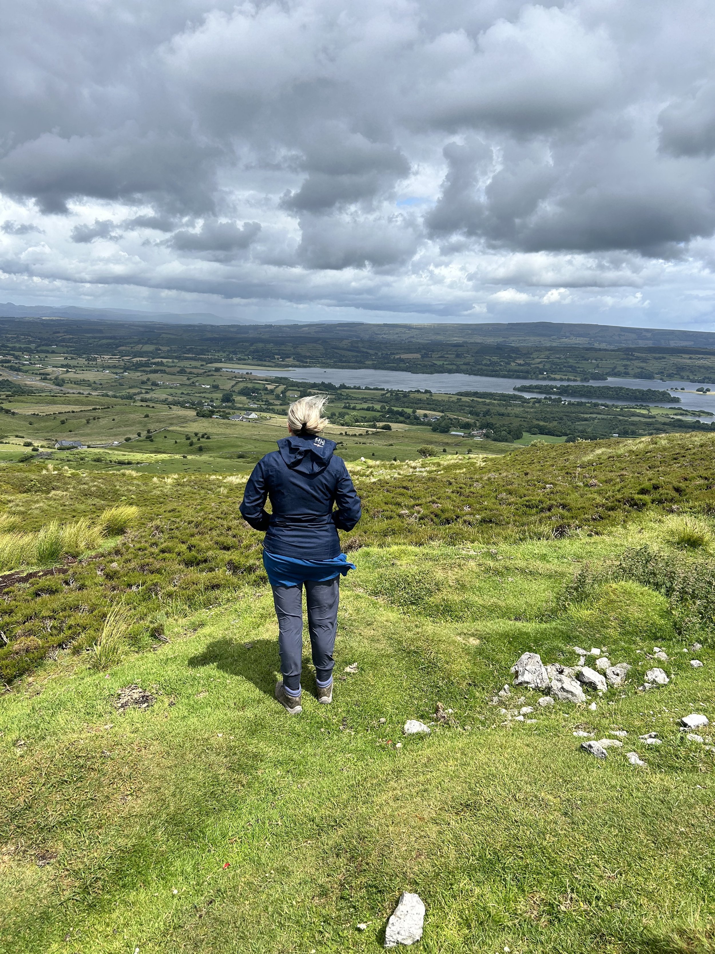 Person standing on a grassy hill overlooking a scenic landscape, with cloudy sky and distant lake and hills.