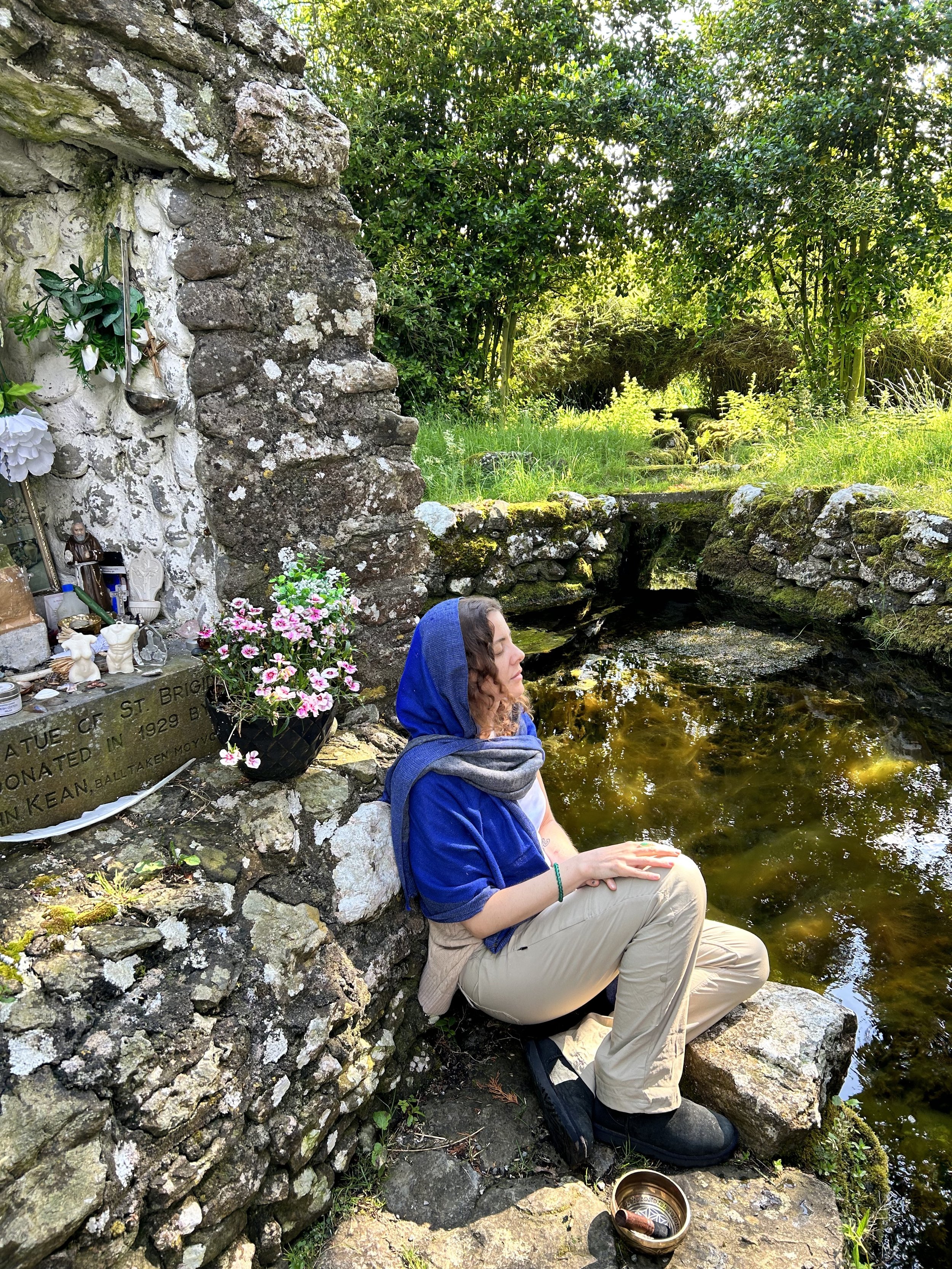 A person wearing a blue hooded outfit sitting by a pond next to a small stone shrine adorned with flowers and religious statues, surrounded by a lush green landscape.