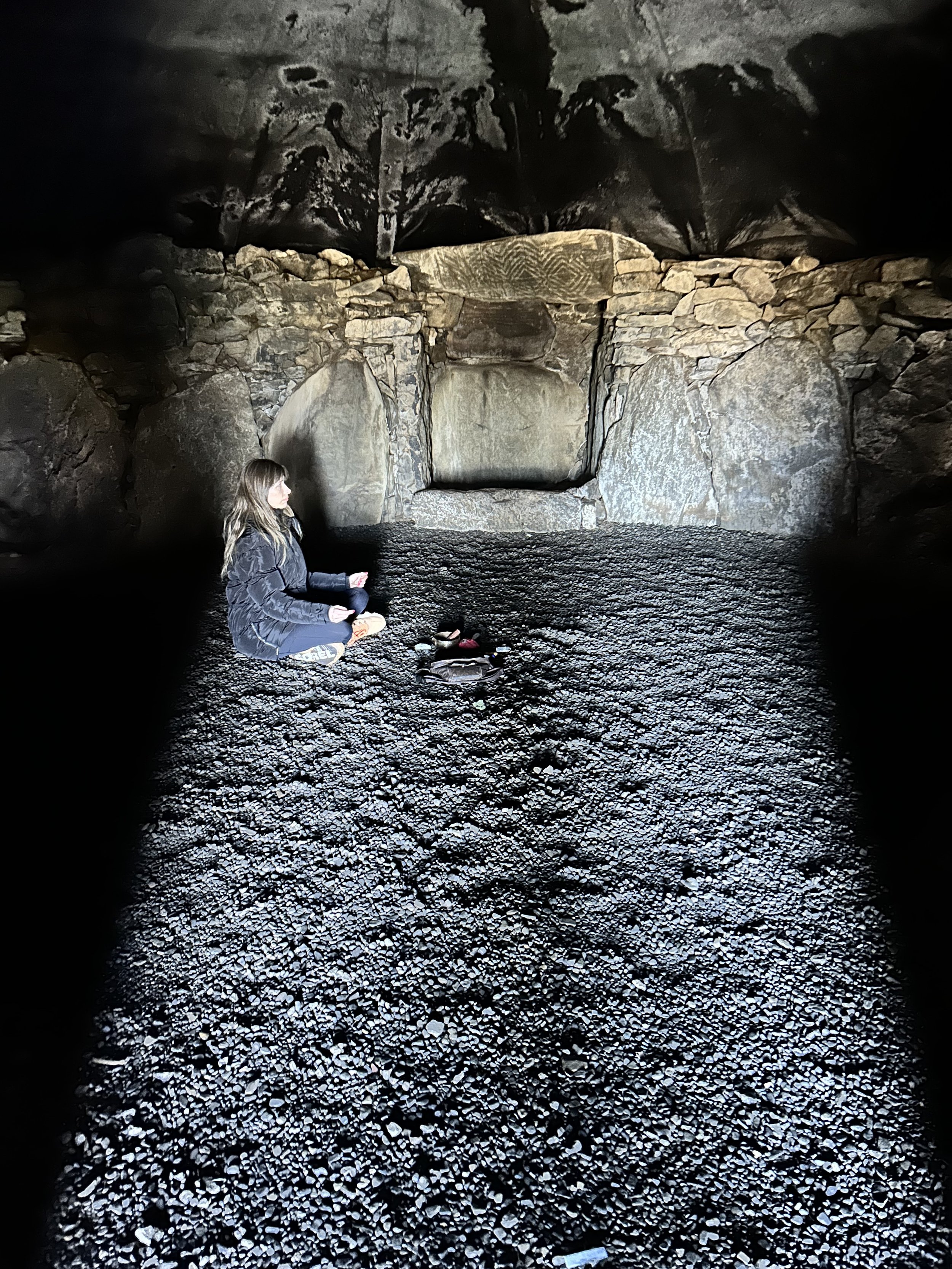 Person sitting cross-legged on a gravel floor inside a stone chamber with rough-hewn walls, partially lit from above.