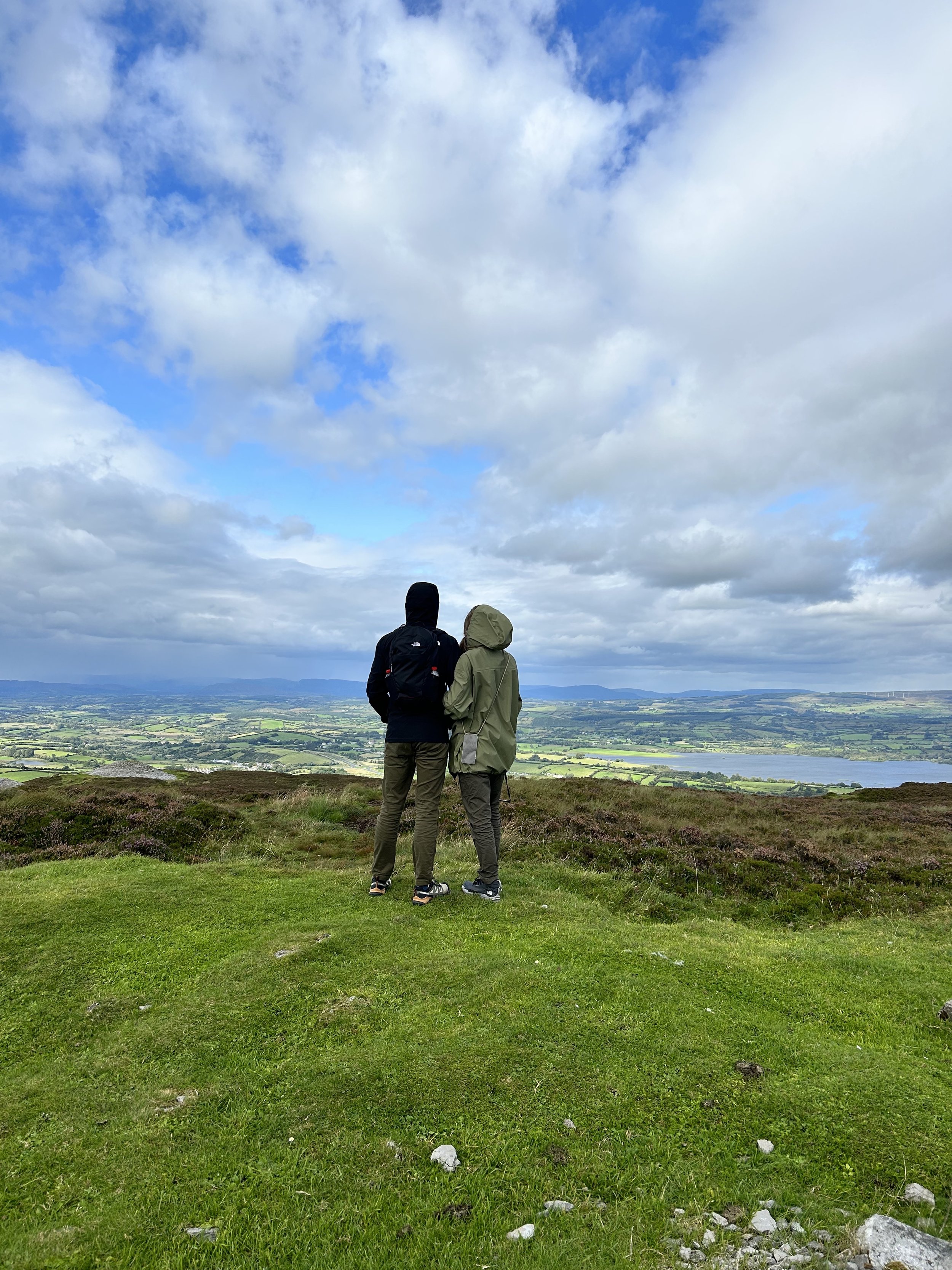 Two people in jackets standing on a grassy hill overlooking a scenic landscape with mountains, valleys, and scattered clouds in the sky.