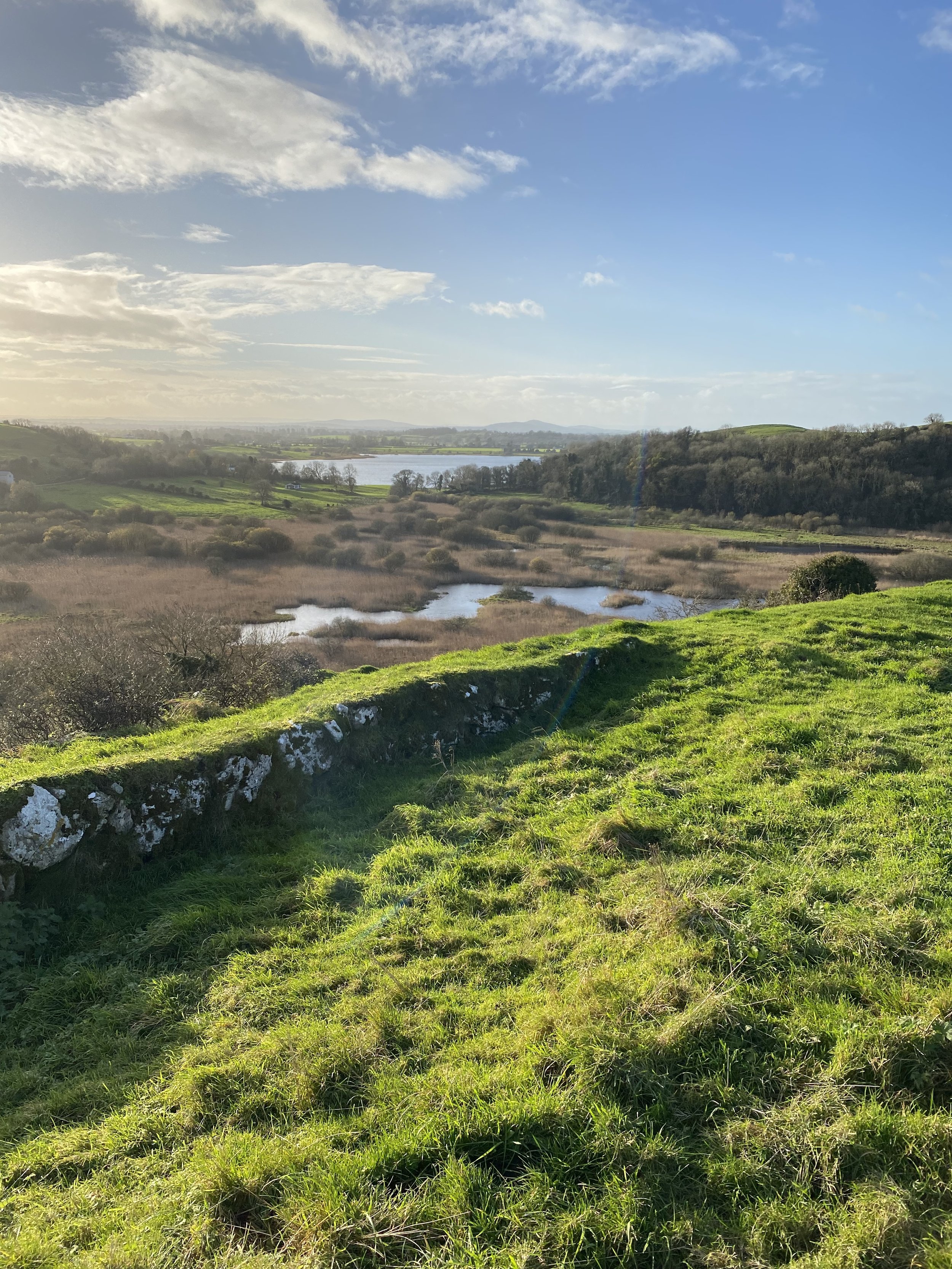 Scenic landscape with green grass, stone wall, wetlands, trees in the distance, and a blue sky with clouds.