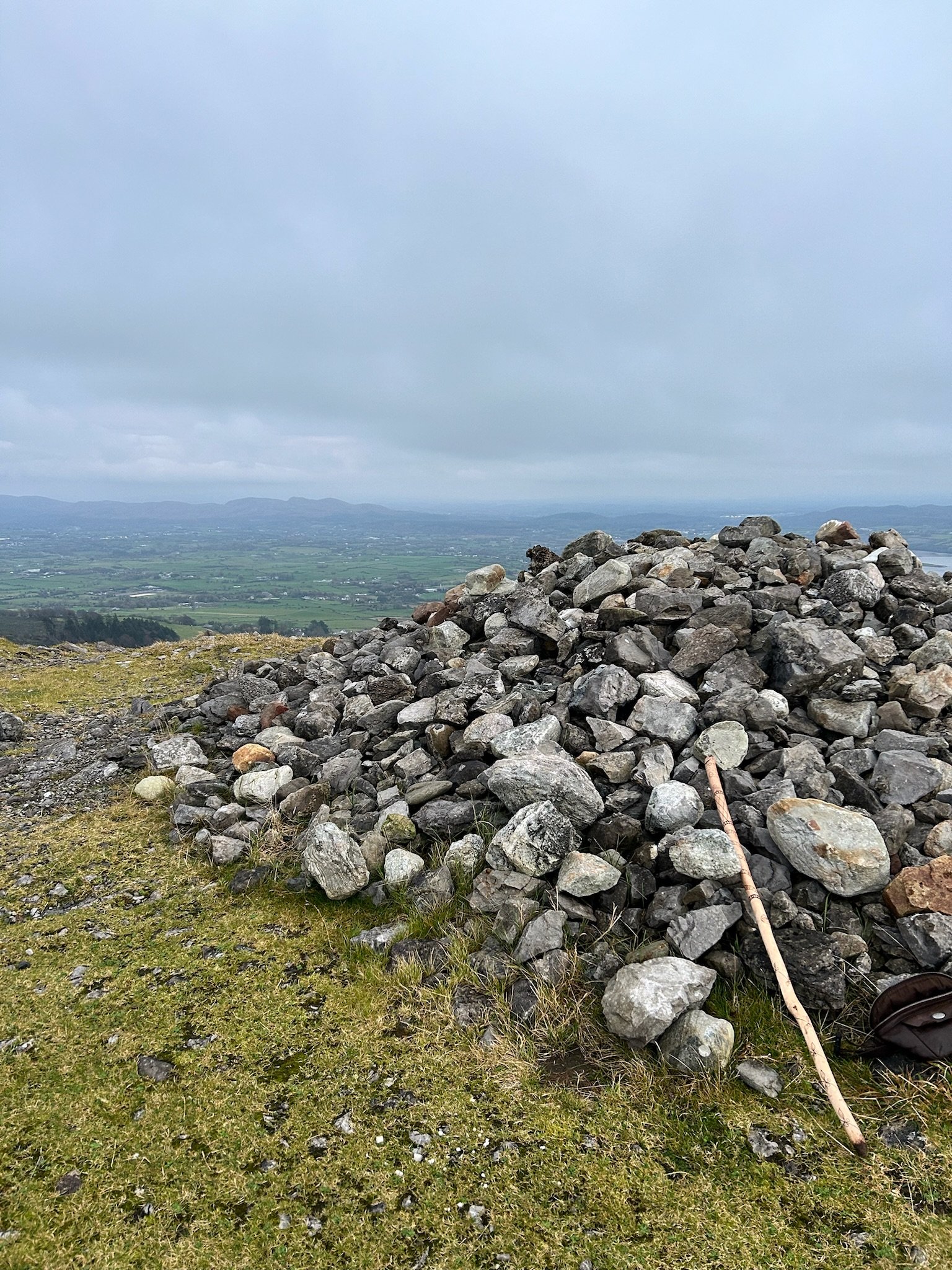 Pile of rocks on grassy hilltop with stick, overlooking rural landscape and hills in the distance.
