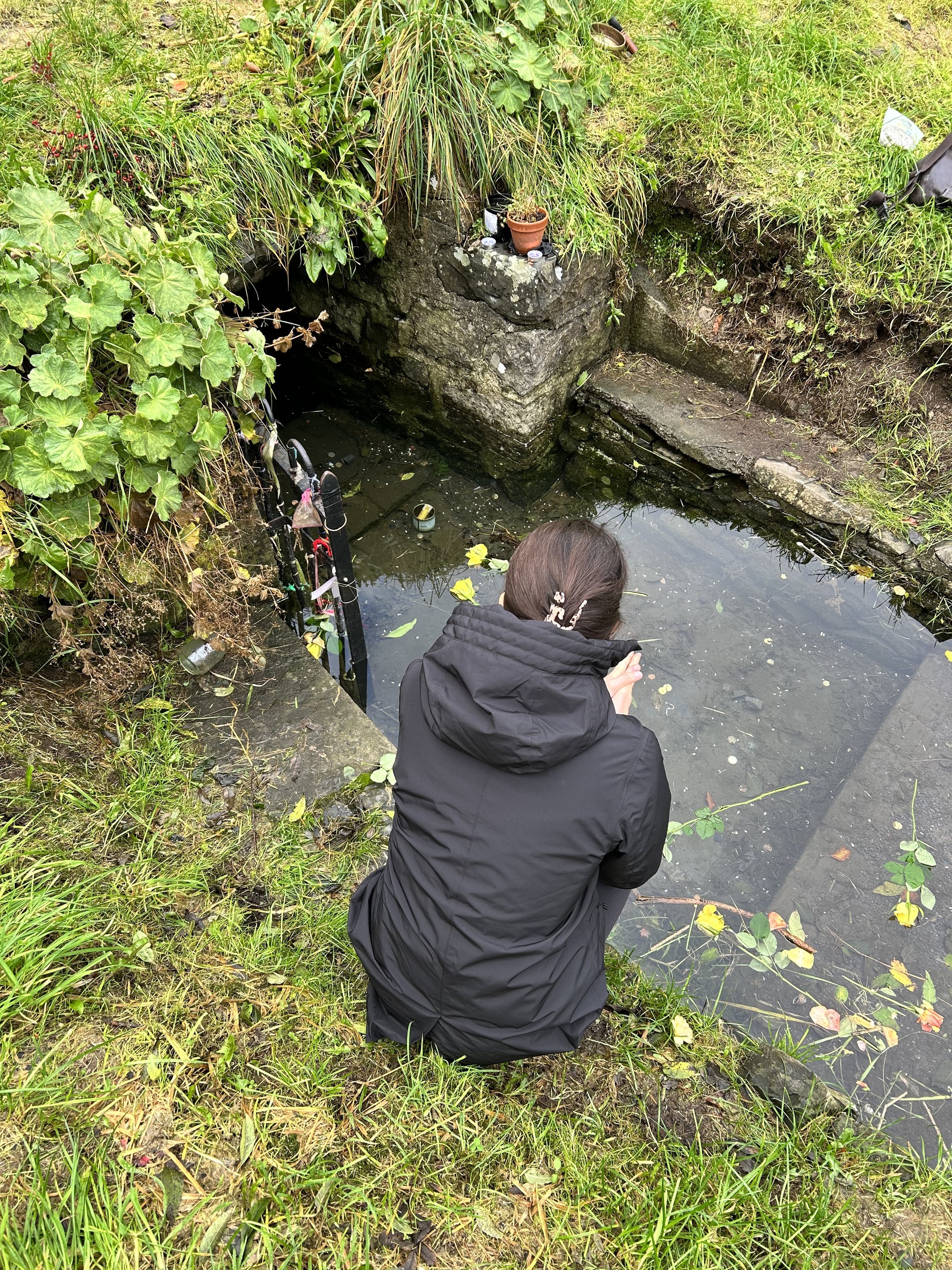 Person in a black coat kneeling by a small outdoor pool surrounded by greenery, with offerings and candles placed nearby.