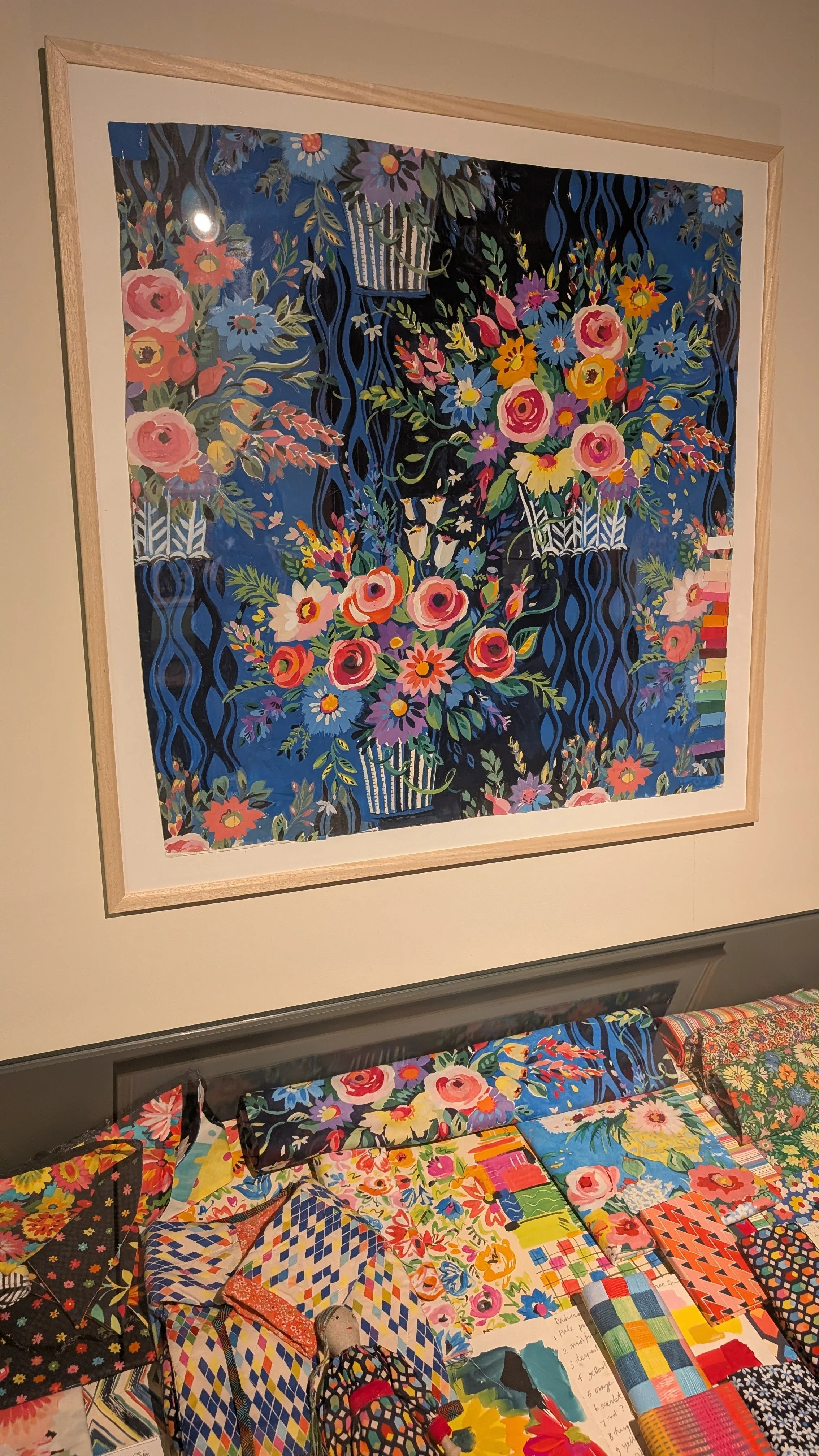 Framed flower painting on the wall and colourful floral fabric samples on the table below in the Sarah Campbell exhibition