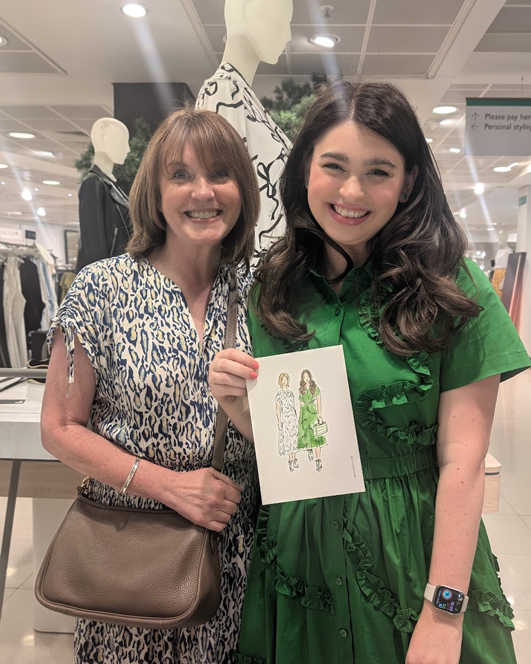 Two women stood in a department store smiling and holding a custom portrait of themselves illustrated by Helen Ridley. One lady wears a patterned blouse and the other wears a green dress.