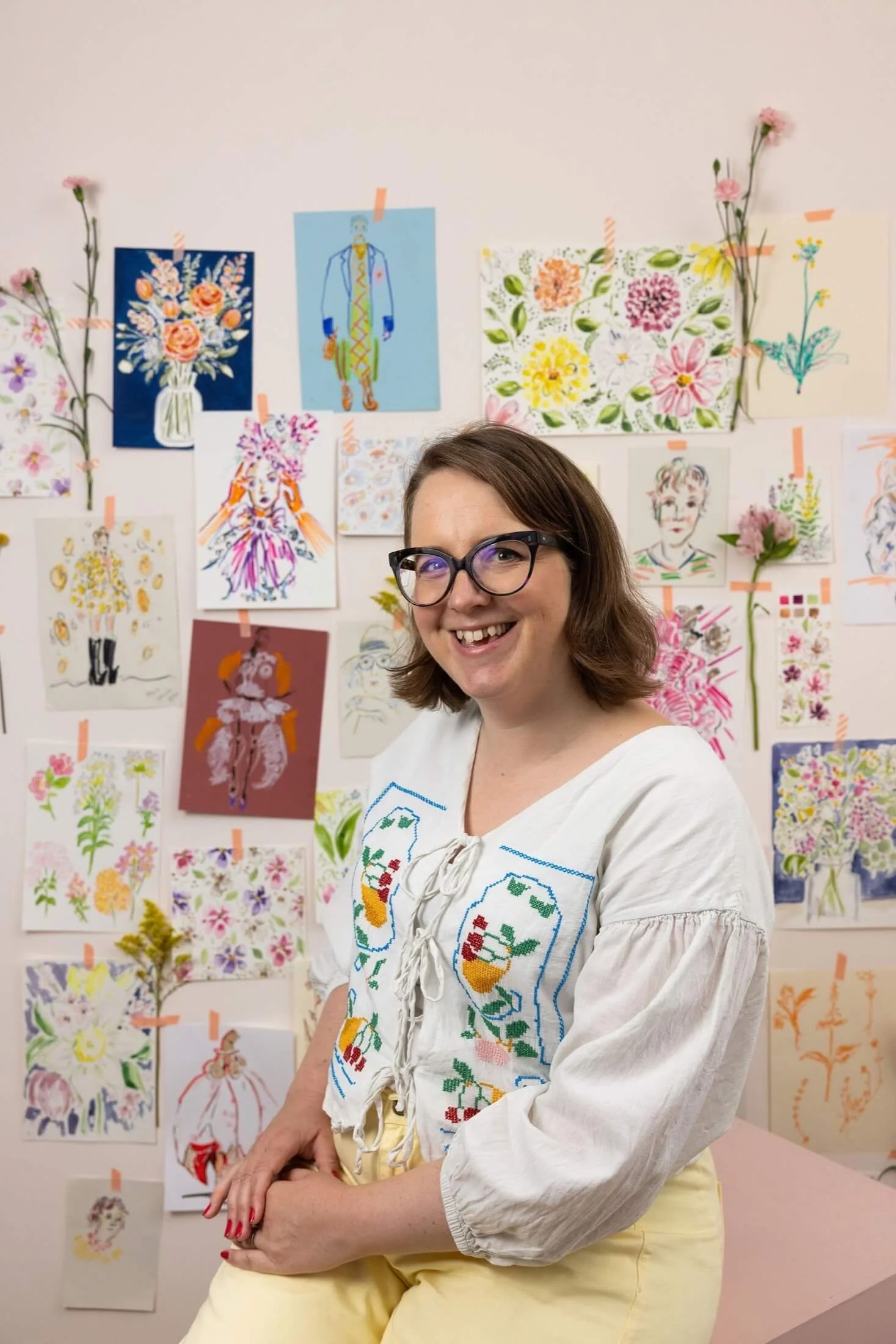 Helen Ridley smiling at the camera, sat in front of a wall covered in watercolour illustrations and florals