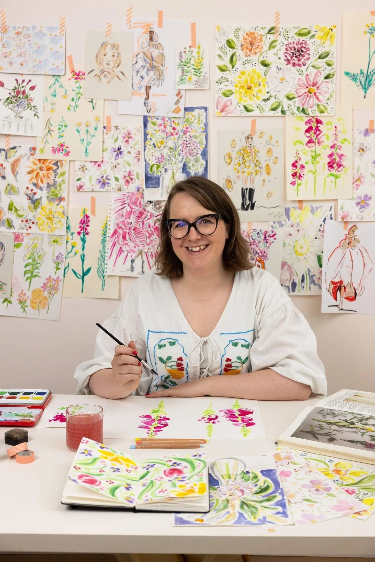 Helen Ridley smiles at the camera sat at a table holding a paintbrush, with a wall of illustrations behind her