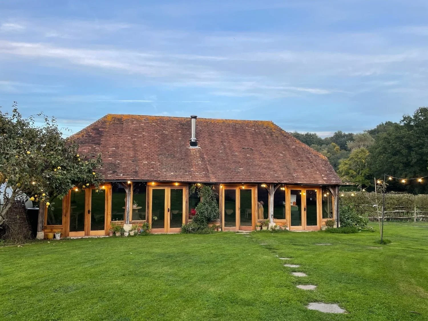 A barn with a red-tiled roof and wooden framing, decorated with string lights, is situated in a grassy yard with a stepping stone path, a lemon tree on the left, and a fenced garden with trees in the background under a blue sky.