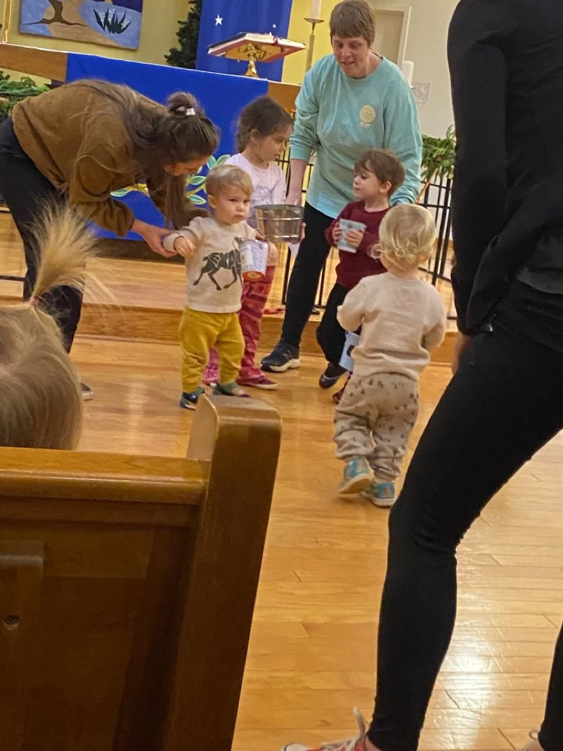 Children and adults participating in a group activity inside a room with wooden floors and colorful decorations, including a blue backdrop and a podium in the background.