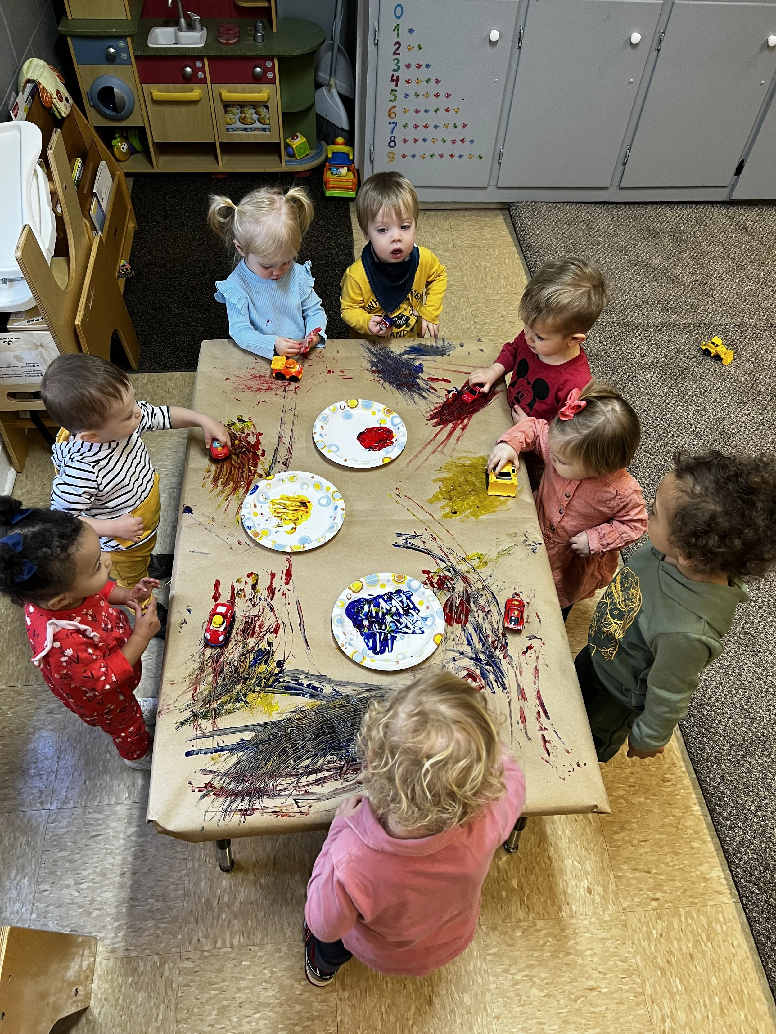Children gathered around a table painting with finger paints and toy cars on a brown paper-covered table in a preschool classroom.
