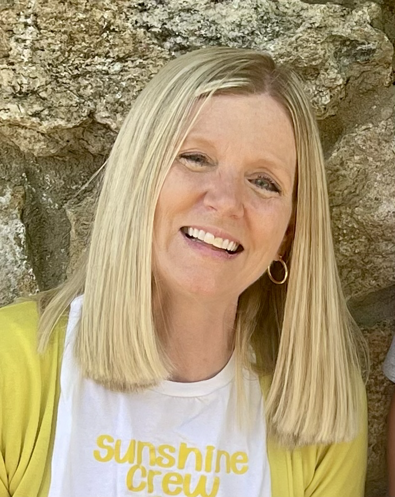 A woman with blonde hair, wearing a white and black striped sweater, smiling, standing in front of a plain light gray background.