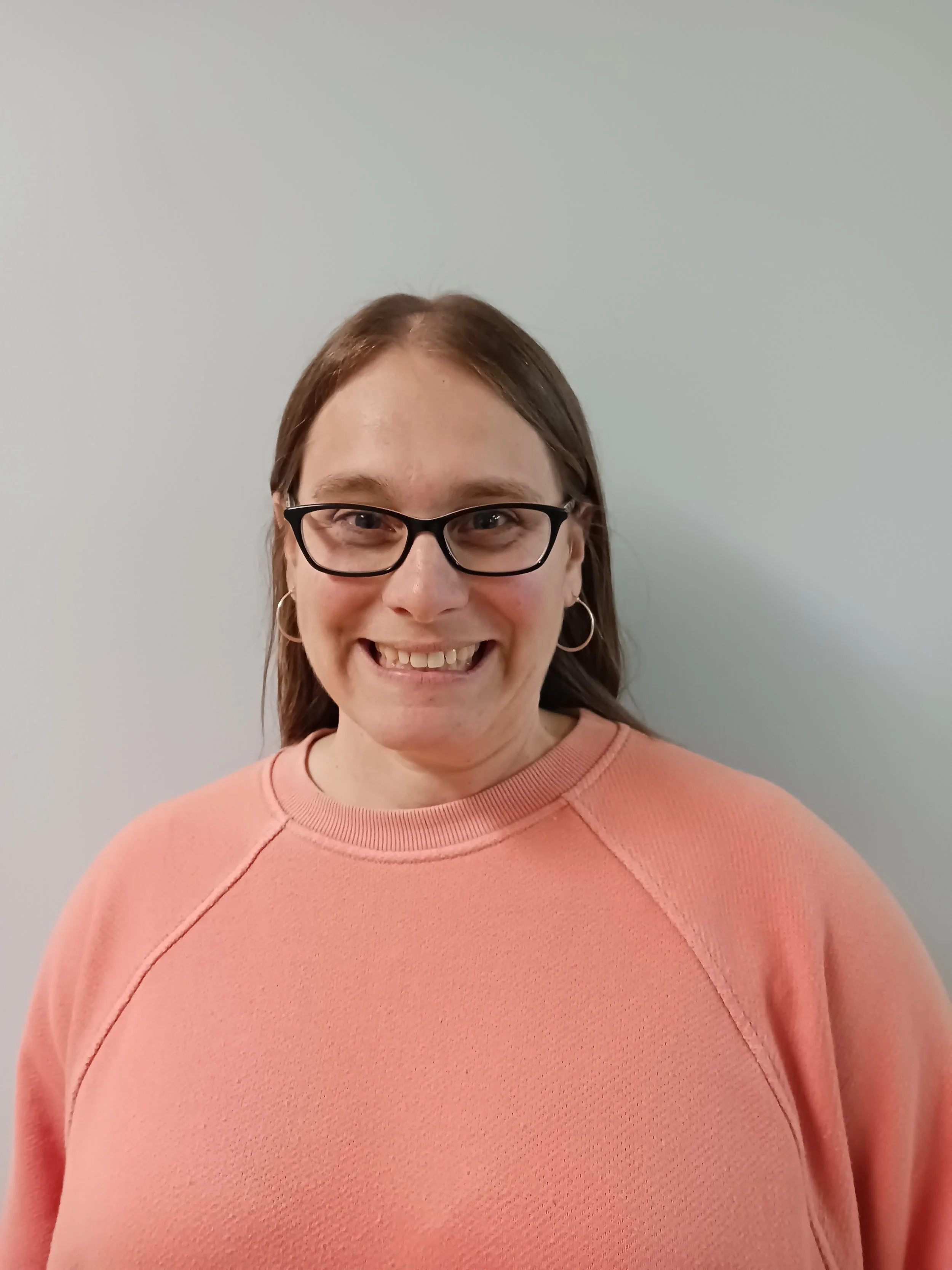 A woman with glasses and hoop earrings smiling, wearing a peach-colored sweatshirt, standing against a plain light gray wall.