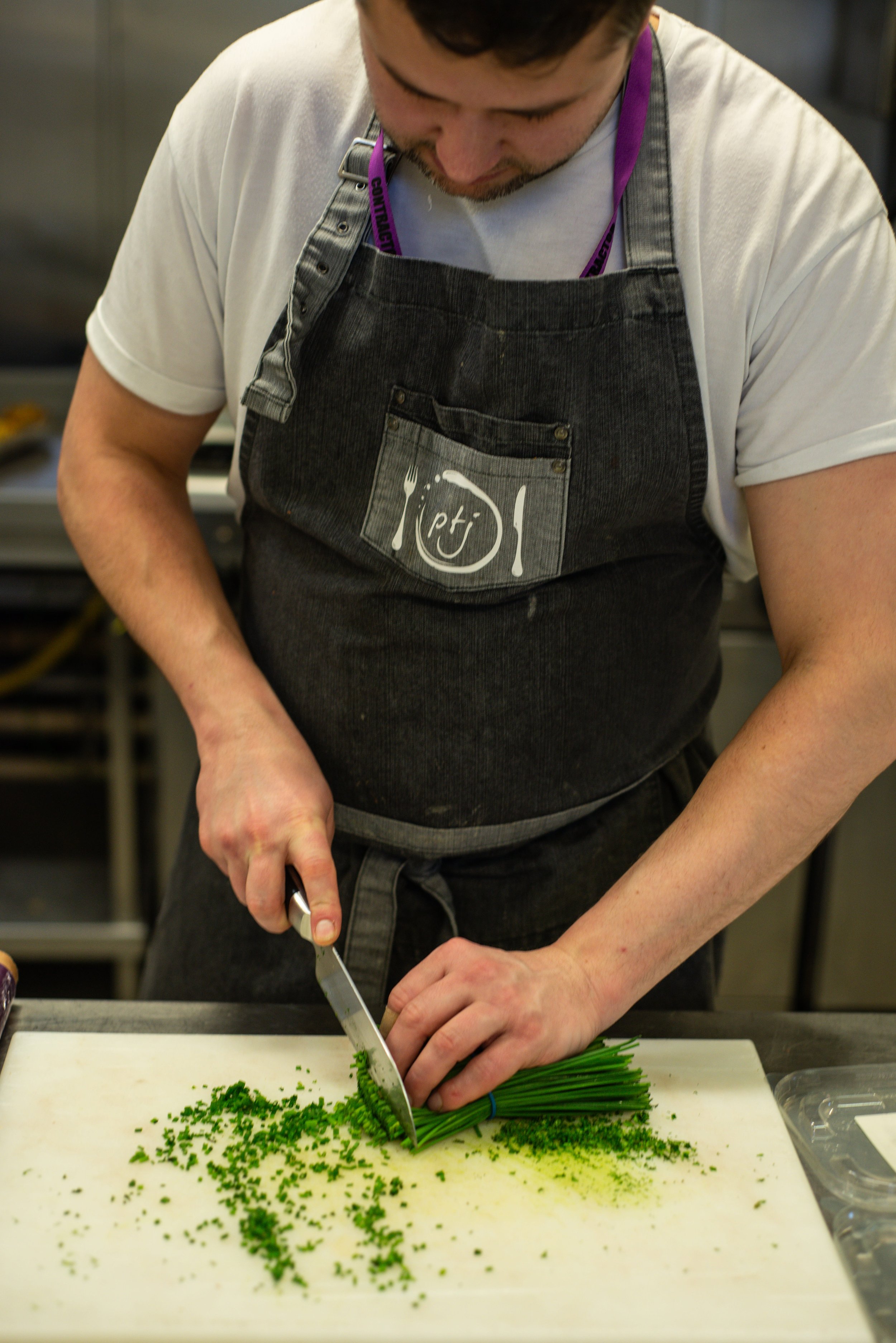 A man wearing a black apron and a white t-shirt chopping chives on a white cutting board in a kitchen.