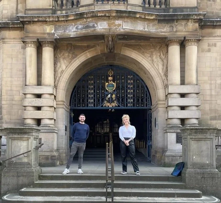 Two people standing on steps in front of an ornate historic building with an arched entrance and iron gates.