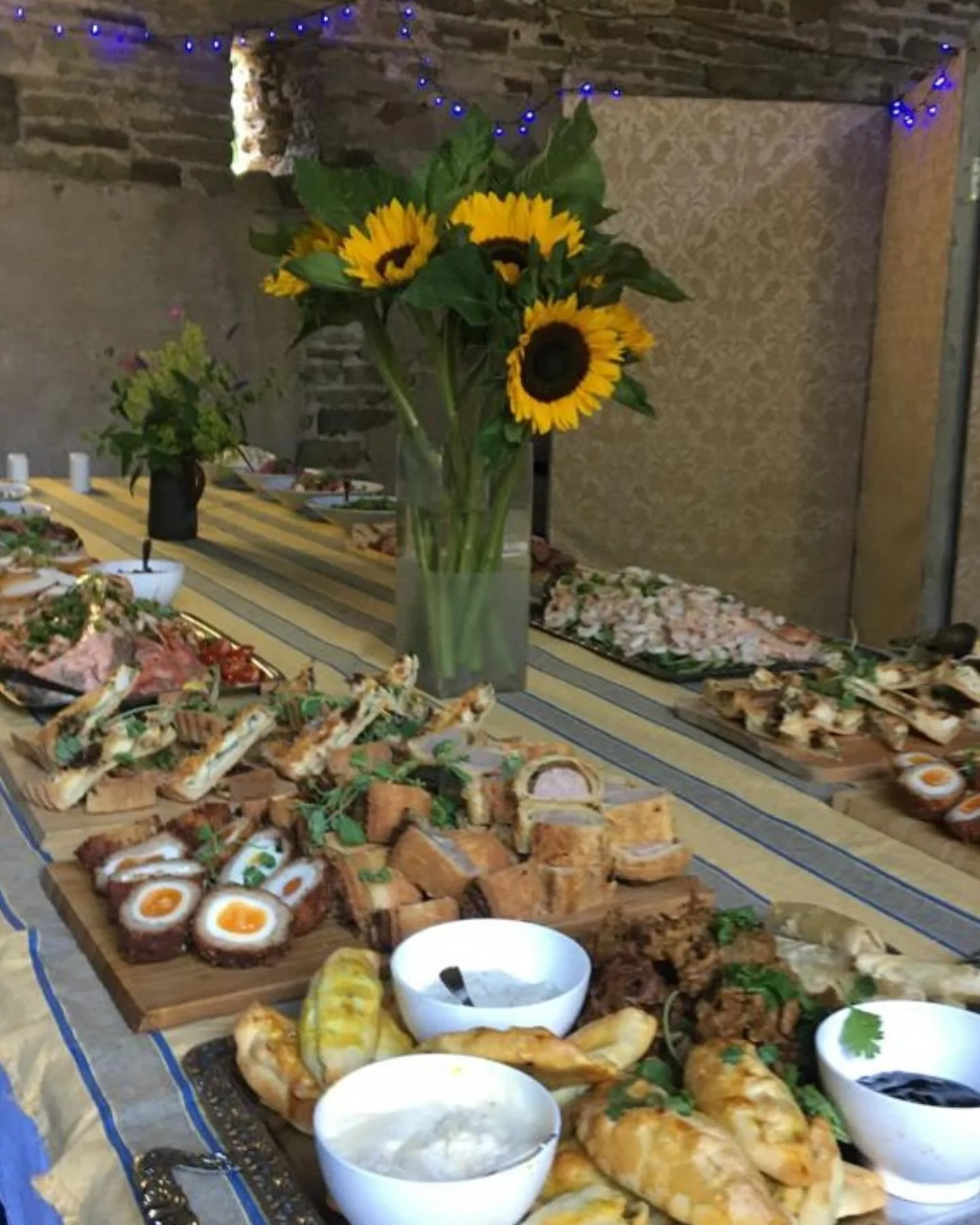 A table set with a variety of appetizers and dips, with a large bouquet of sunflowers in the center, decorated with blue string lights in the background.