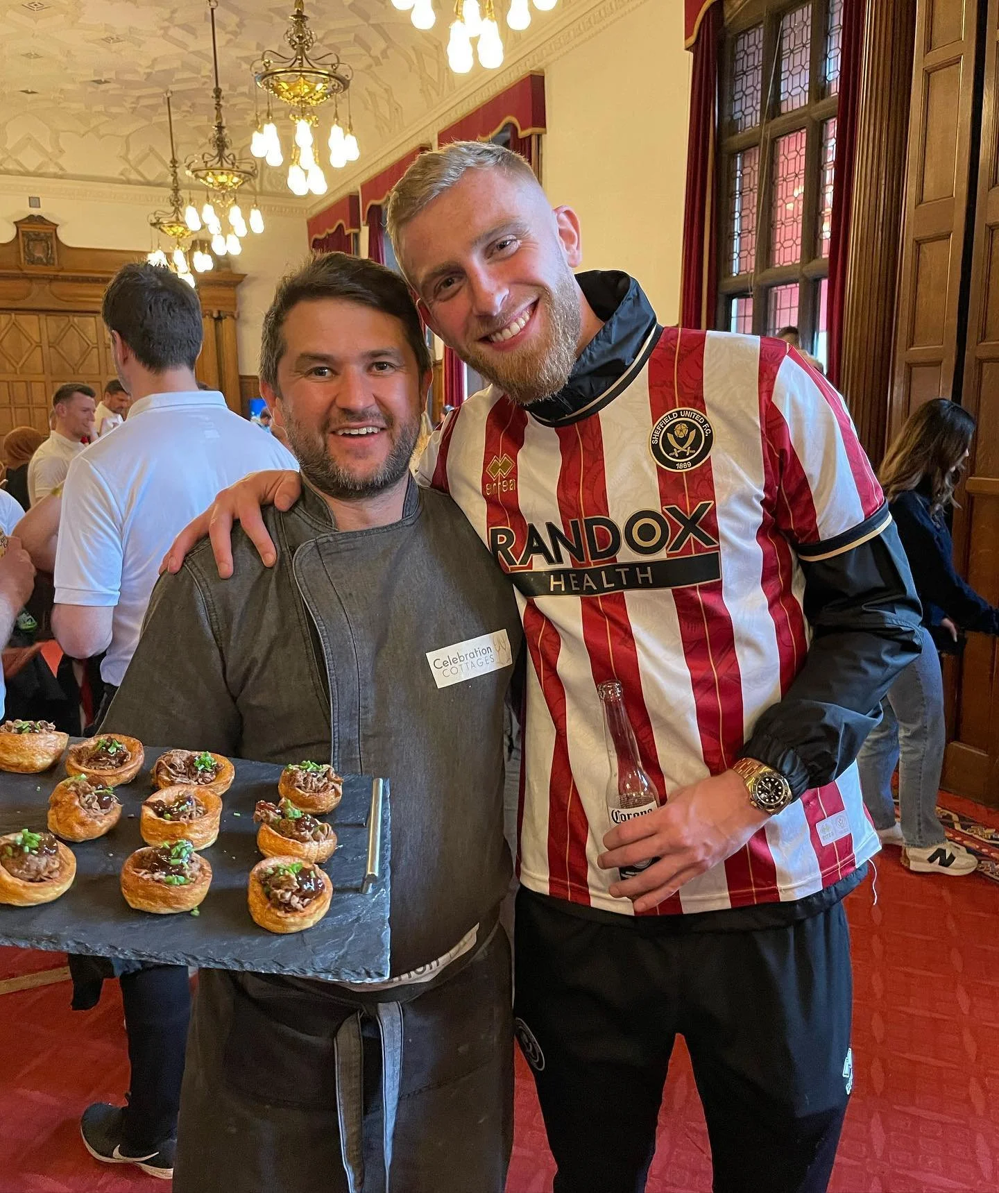 Two smiling men at a social event. The man on the left wears a gray chef jacket and holds a tray of mini appetizers. The man on the right wears a red and white striped soccer jersey and holds a beer bottle. The background features wood-paneled walls, stained glass windows, and other guests.