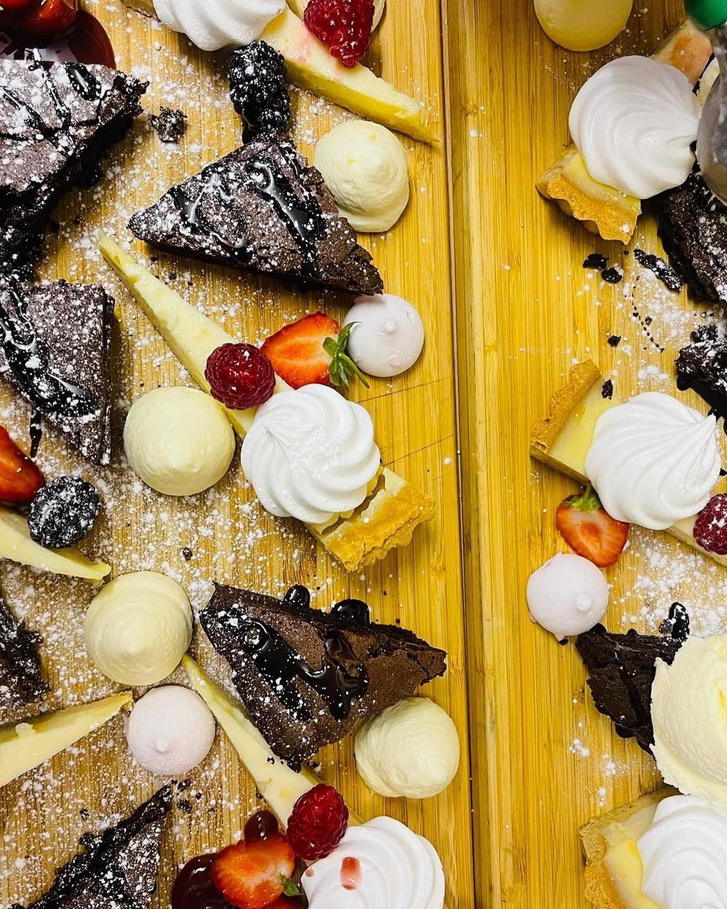 Assorted desserts on a wooden surface, including chocolate brownies, whipped cream, strawberries, raspberries, cream scoops, and lemon slices, topped with powdered sugar and chocolate drizzle.