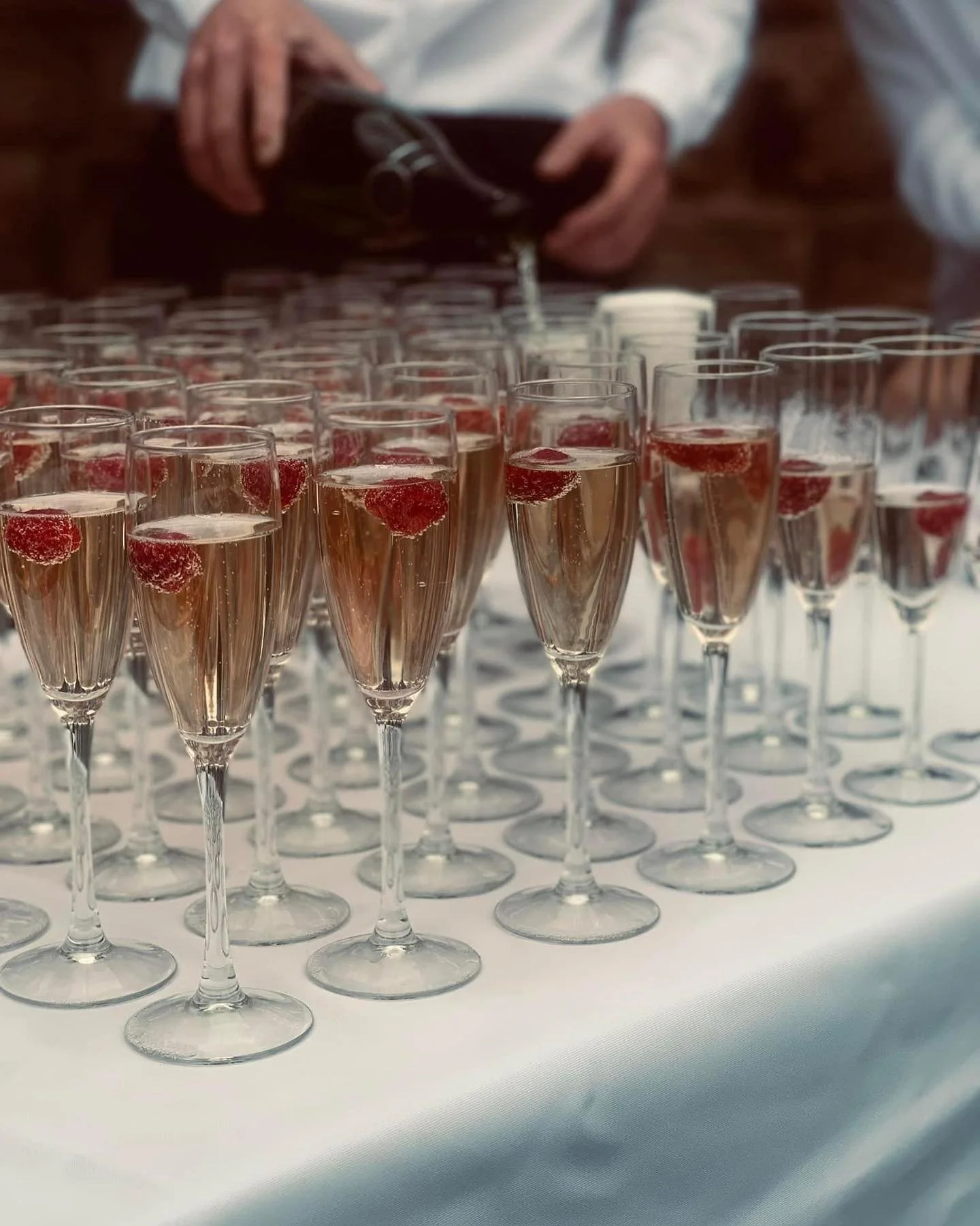 Multiple glasses of pink champagne with raspberries on top arranged on a white tablecloth, with a person in the background pouring more champagne from a bottle.