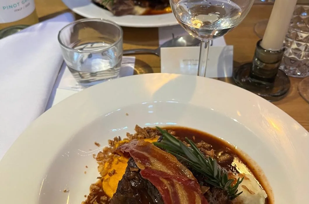 Plate of gourmet dish with meat, bacon, herbs, and sauce on a white plate, with wine glass, water glass, and candles on the table.