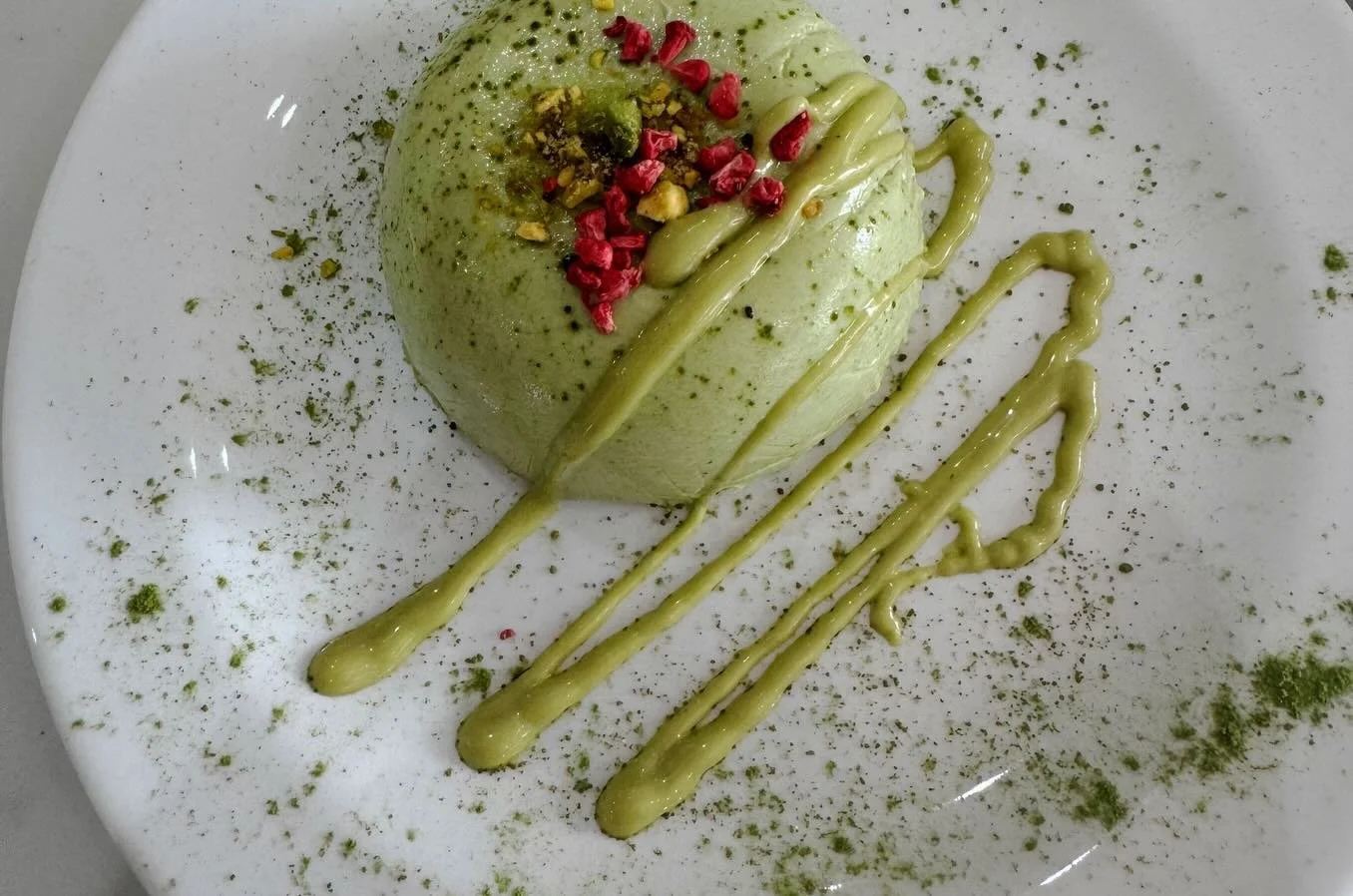A green mousse dessert garnished with chopped nuts and dried berries, decorated with green sauce and green powder on a white plate.