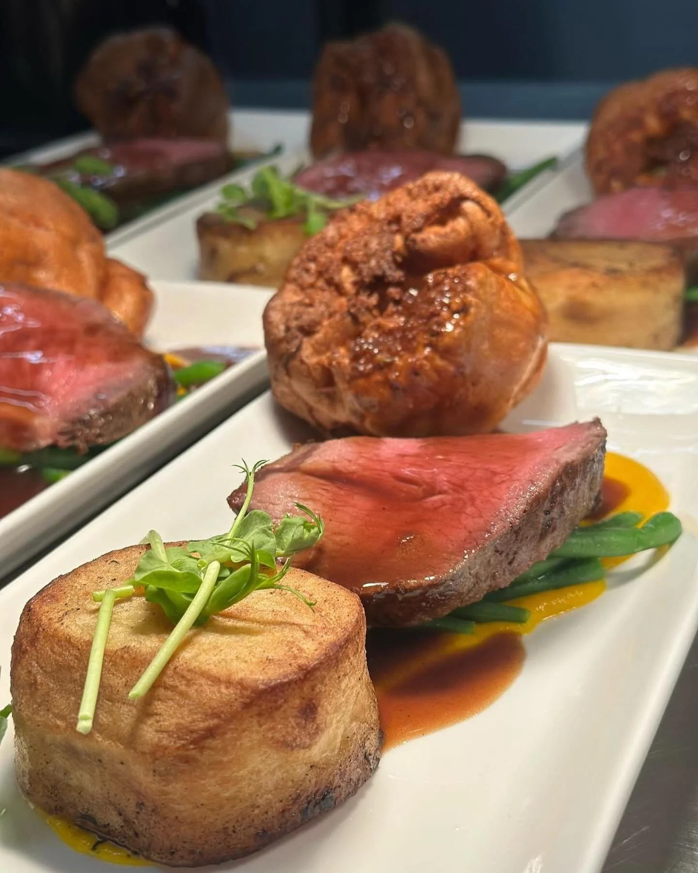 A plated meal featuring a piece of medium-rare steak, a crispy potato cake garnished with microgreens, and roasted or fried meatballs with a garnish of green beans and sauce.