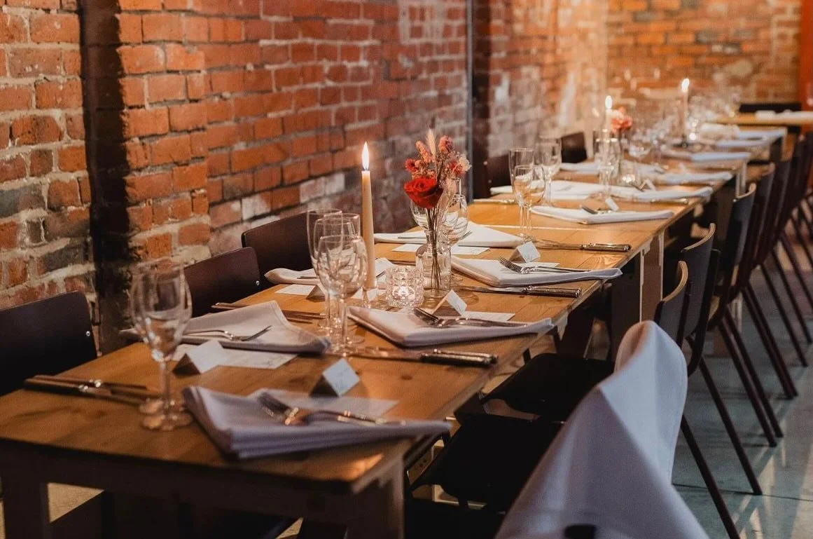 A long dining table set for a special occasion, with candles, floral centerpieces, wine glasses, and white napkins in a brick-walled restaurant or event space.