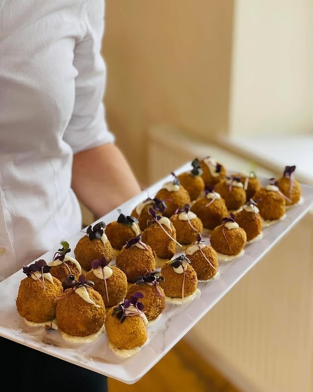 A person holding a large white rectangular platter with multiple breaded and deep-fried appetizer balls garnished with microgreens and a drizzle of sauce.