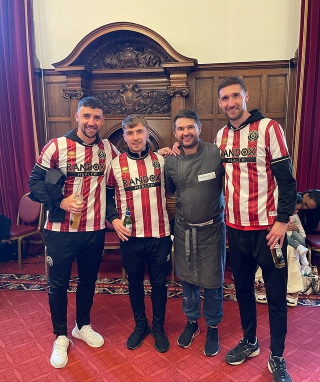 Four men standing together in front of a wooden ornate wall, three of them wearing matching red and white striped sports jerseys, and one in a gray chef's apron, smiling and holding drinks.