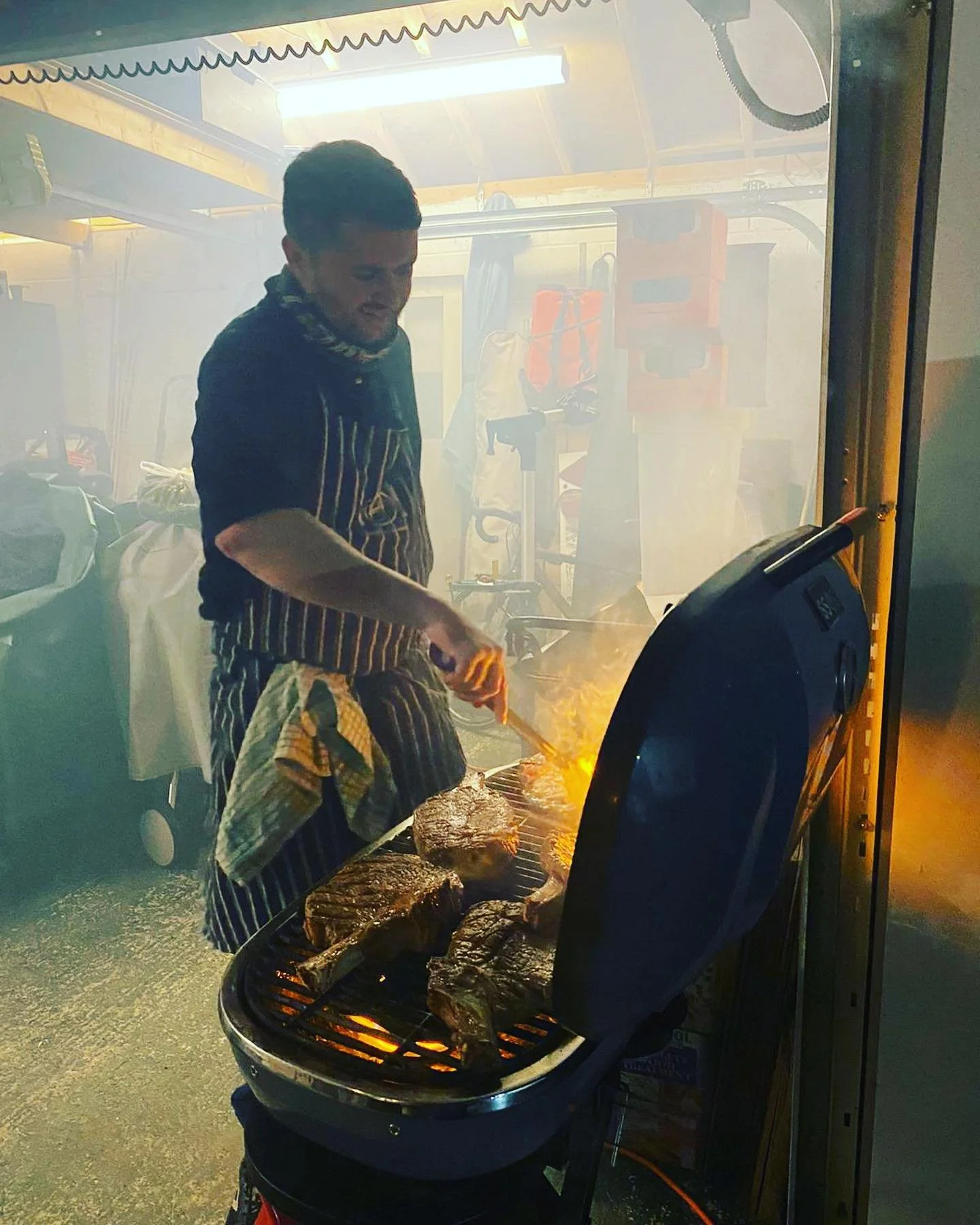 Man cooking steaks on a grill in a garage or workshop, using tongs, with smoke and flames present.