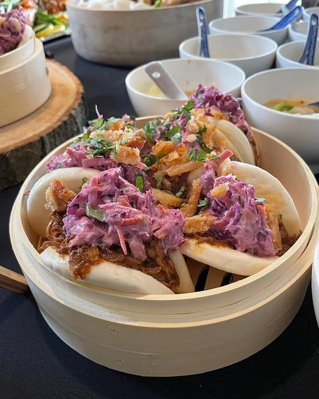 Three bao buns filled with shredded pork, purple coleslaw, and fried shallots in a bamboo steamer basket.