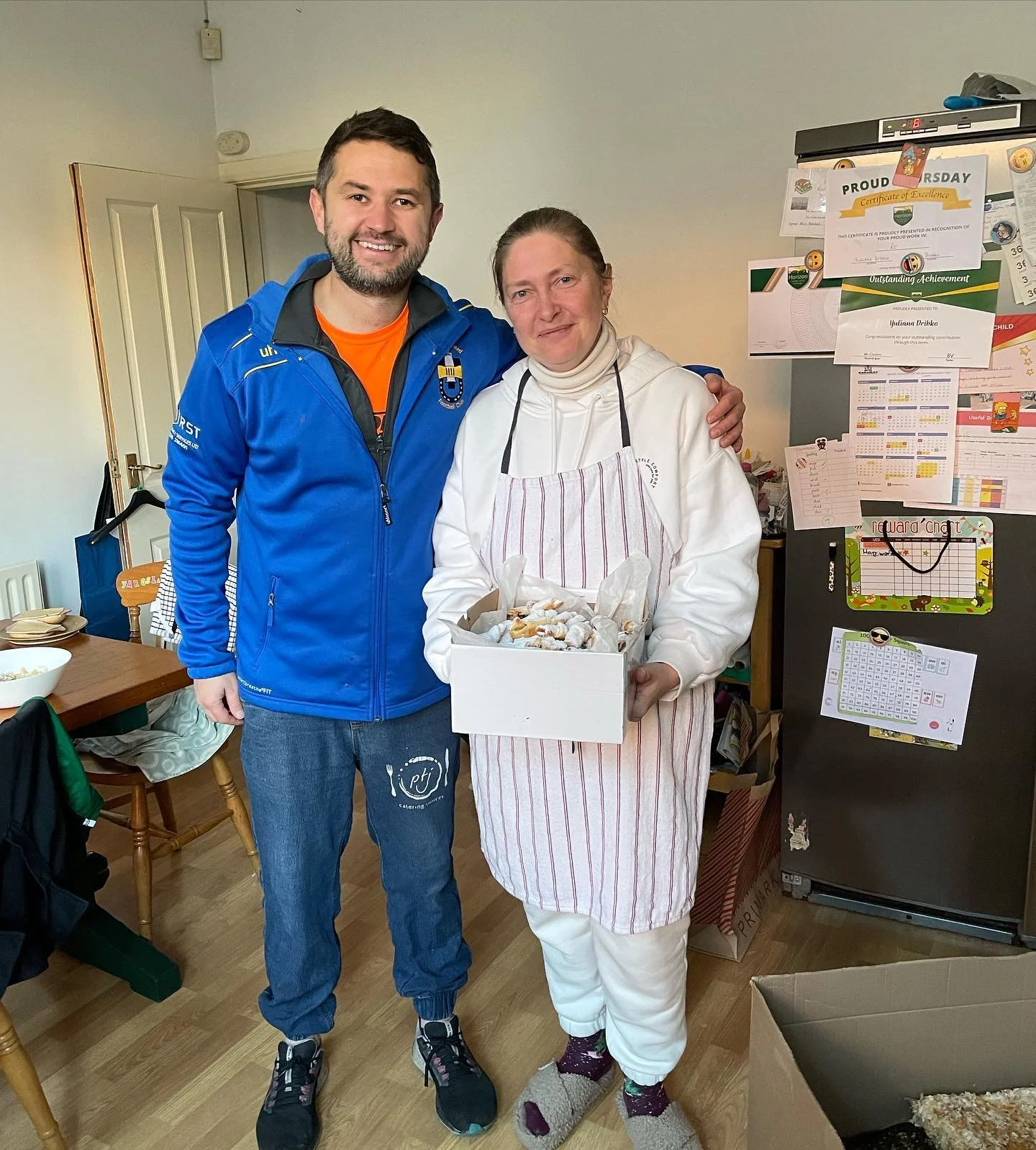 A man and woman stand together indoors, smiling at the camera. The woman is holding a box of cookies or baked goods. The background includes a refrigerator with photos and notes, and a dining area with chairs and a table.