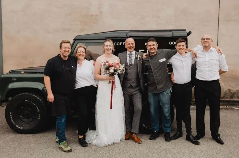 Group of seven people including a bride in a white wedding dress holding a bouquet, standing in front of a black van, smiling and posing together outside.