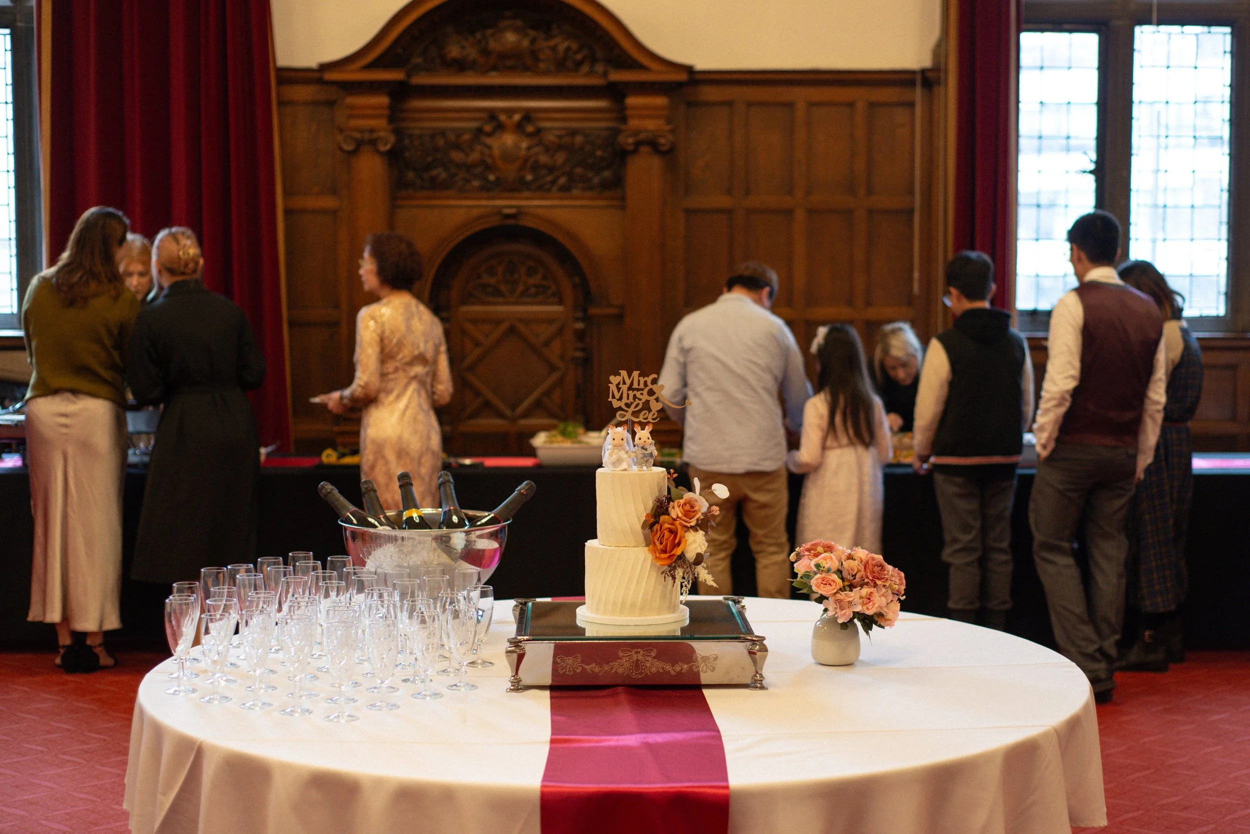 A wedding reception with a white cake decorated with flowers and a topper that reads 'Mr & Mrs Lee', a bouquet of pink flowers, glasses, and bottles of champagne on a round table, and guests in formal attire gathering in the background near a buffet table in a wood-paneled room with large windows.
