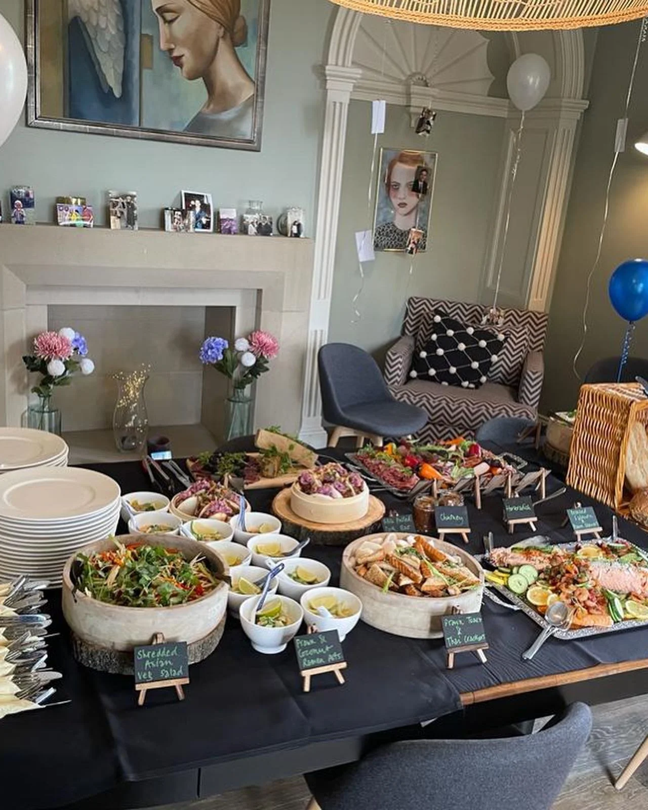 A buffet table with various dishes, salads, and appetizers set up for a gathering in a decorated living room with artwork, balloons, and flowers.