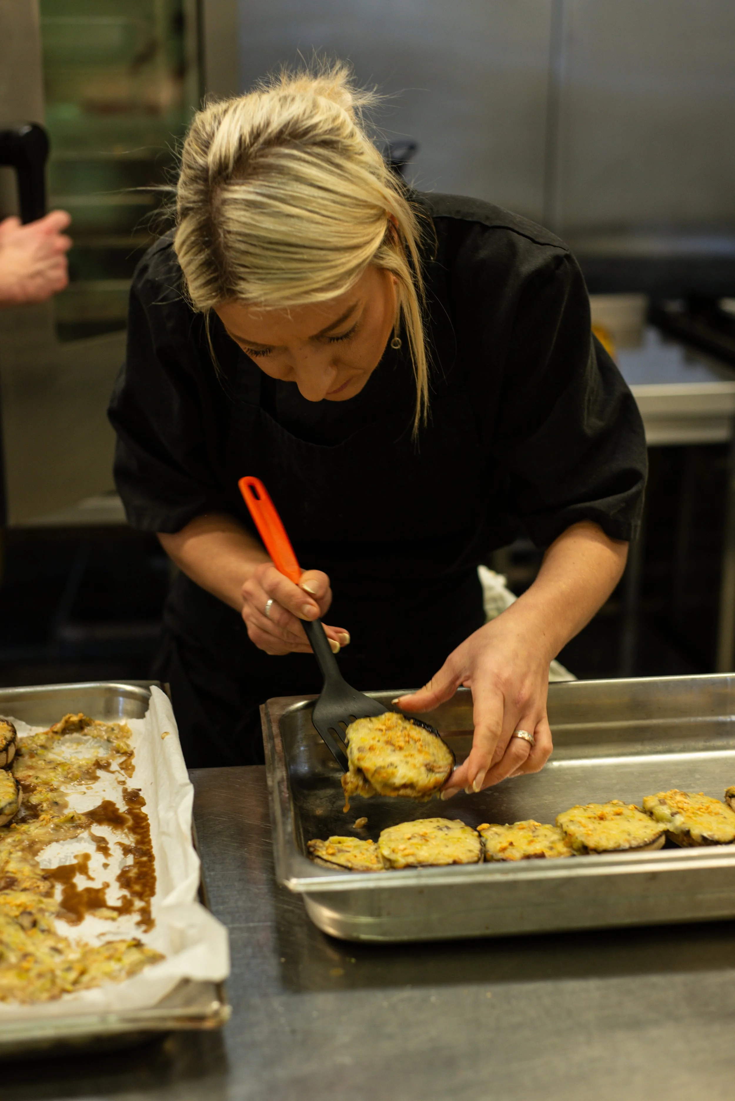 A woman with blonde hair wearing a black apron is placing a baked eggplant dish onto a tray in a commercial kitchen.